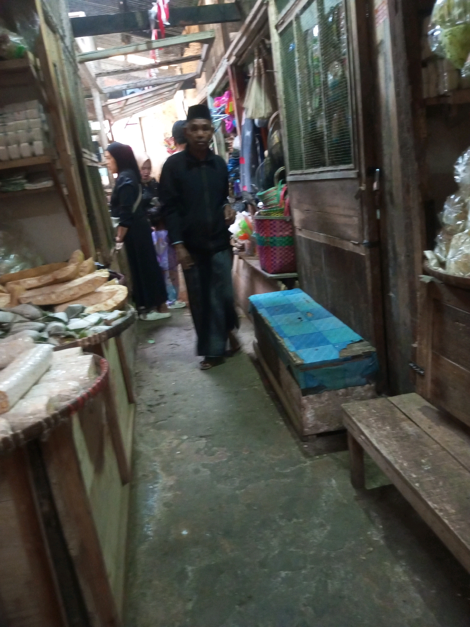 The image captures a narrow marketplace aisle in Kalibeber, Indonesia. A man wearing a black cap and dark clothing walks towards the viewer. To his left, a woman in a black hijab and dress is partially visible, facing away from the camera. Another person, possibly a woman in a lighter colored hijab, is behind her. The aisle is lined with stalls constructed from wood. On the left, shelves are stocked with white items, and a display of what appears to be dried fish or wood is in the foreground. On the right, a wooden structure with a checkered blue tarp-covered top serves as a bench or seating area. A wooden bench is also present in the far right of the image. The ground is a rough, concrete surface. The overall impression is of a busy, somewhat dimly lit marketplace.
