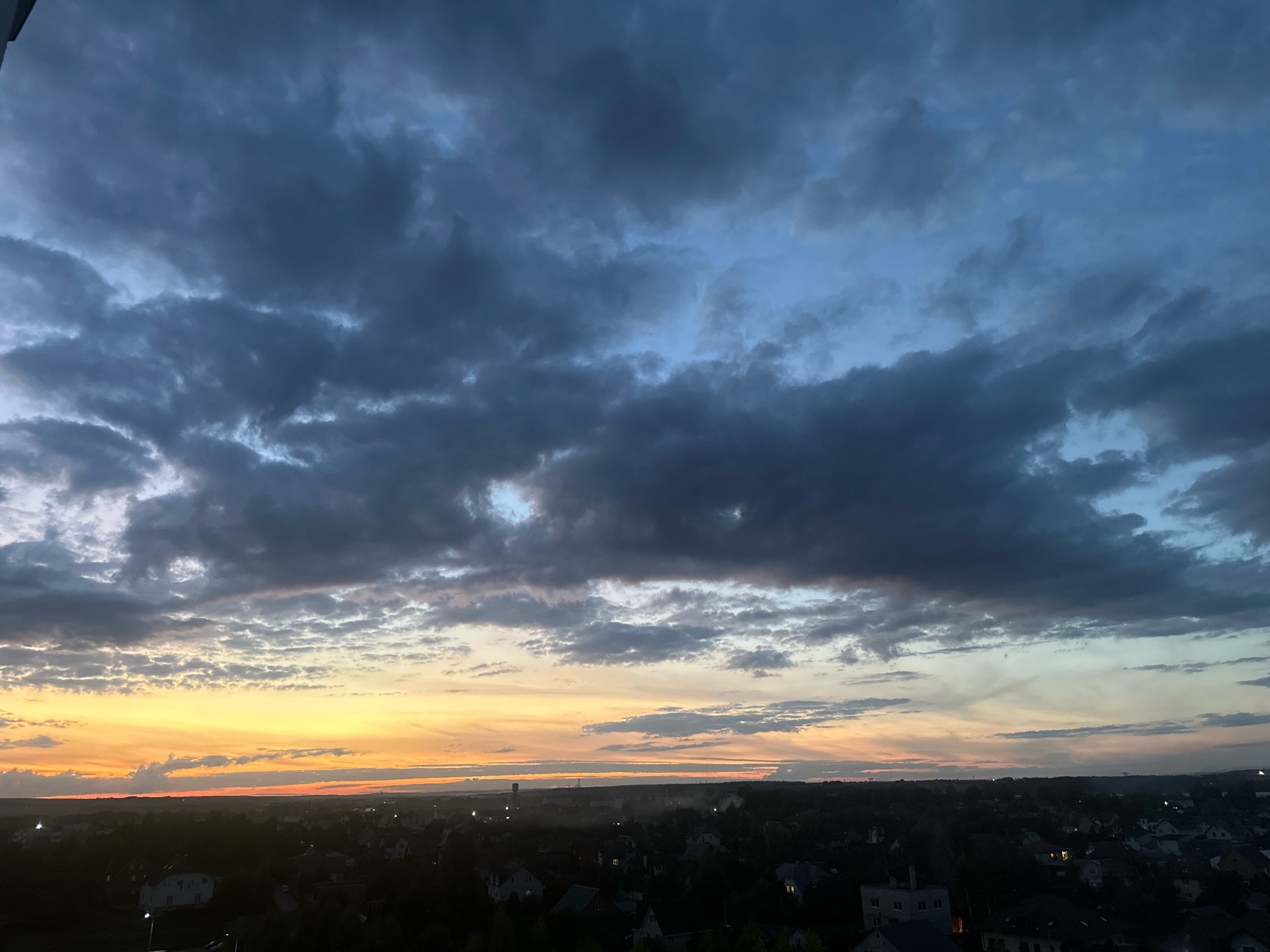 The image captures a panoramic view of the sky and a cityscape at dusk. The sky is dominated by dramatic, dark gray clouds that stretch across the upper half of the frame. Patches of lighter blue sky are visible between the clouds, particularly towards the right side. A vibrant sunset is occurring on the horizon, with bands of orange, yellow, and a hint of red painting the sky just above the landscape.
Below the sky, a silhouetted cityscape is spread out. Numerous houses with pitched roofs and some larger buildings are visible, their forms dark against the fading light. The lights are on in many of the houses, creating small points of illumination. Trees are also scattered throughout the landscape, adding to the overall dark silhouette. The scene suggests a residential area on the outskirts of a settlement.
Based on the context provided, this image is located in the vicinity of Шчомысліцкі сельскі Савет, Belarus. The image does not contain any discernible people, specific actions, or interactions. The overall impression is one of a natural phenomenon (sunset and clouds) juxtaposed with human settlement.
