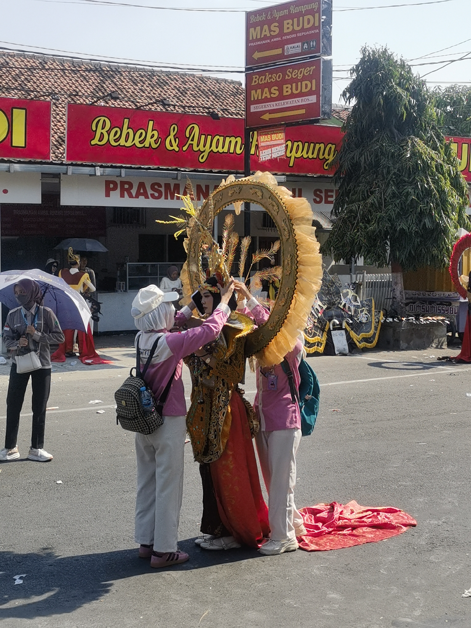 The image depicts a street scene in Blora, Indonesia, possibly during a cultural event or parade. In the foreground, three individuals are clustered together. One person, wearing a traditional elaborate golden headdress and costume with a long red train, is being assisted by two others. The person in the center, wearing a pink jacket, white cap, and light-colored pants, is adjusting the headdress. Another person in a grey jacket and dark pants, holding a purple umbrella, stands to the left, observing. Behind them, several other people are visible, some holding umbrellas and wearing traditional attire.
The background features a building with a red awning and signage for "Bebek & Ayam Kampung MAS BUDI" (Duck & Kampung Chicken MAS BUDI) and "Bakso Seger MAS BUDI" (Fresh Meatball MAS BUDI). A sign pointing to the left indicates "PRASMANAN, AMBIL SENDIRI SEPUASNYA" (Buffet, Take as much as you want). Further in the background, a large, leafy tree stands tall, and various decorations, including what appear to be puppets or artistic props, are visible. The overall setting is outdoors on a paved street under a clear sky.