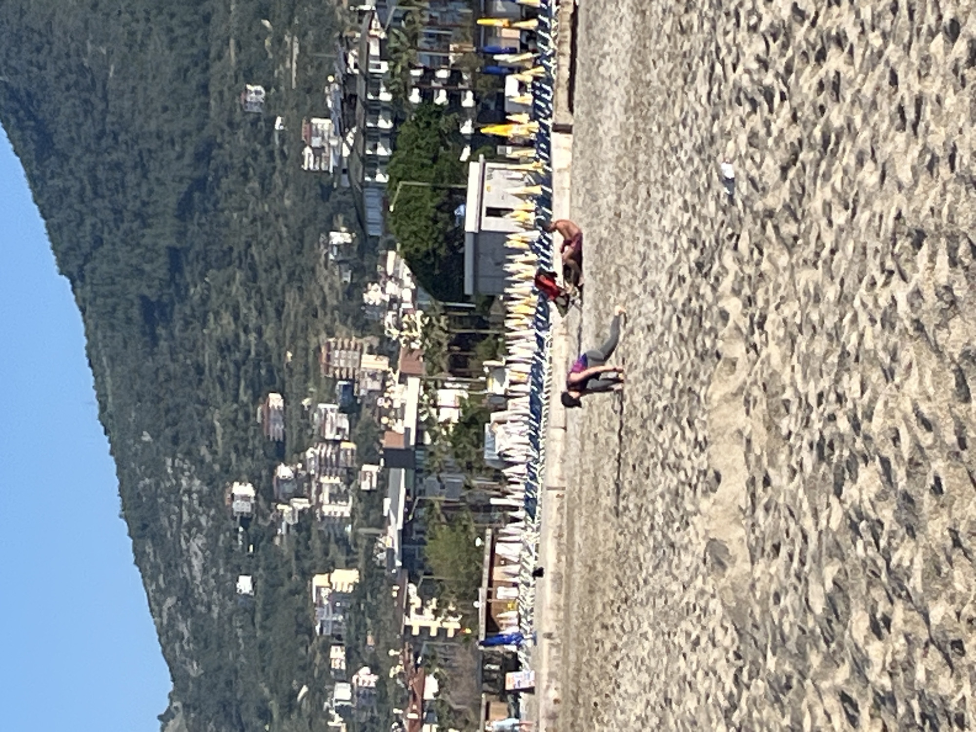 The image captures a beach scene in Alanya, Turkey. The beach, composed of light-colored sand, stretches along the coastline. Rows of beach umbrellas and loungers are positioned close to the water. A man and woman are on the beach. The buildings of Alanya line the coastline, with a backdrop of a mountain covered in dense green vegetation. The sky is a clear blue.