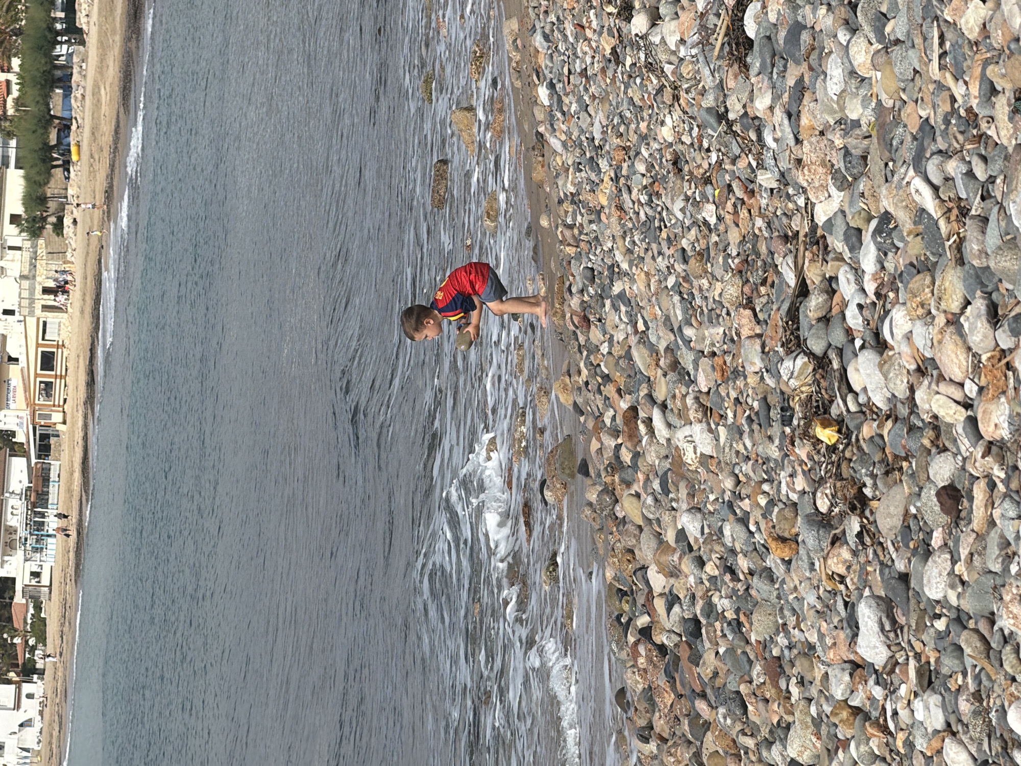 The image depicts a daytime coastal scene on a pebbly beach in Mont-roig del Camp, Spain. The immediate foreground is dominated by numerous rounded stones of varying sizes and colors, including shades of grey, white, brown, and reddish-brown, along with some dried seaweed and debris.
A young male child, estimated to be between three and five years old, stands in a crouching position at the water's edge in the midground. He has short brown hair and is wearing a red and yellow short-sleeved shirt and grey shorts, with bare feet. He is holding a smooth, light-colored, ovoid stone with both hands, his gaze directed towards the water. Small waves are breaking around his feet and against other partially submerged rocks, creating white foam.
The sea extends into the background with a blue-grey hue and visible surface ripples. Across the water, a distant sandy beach is visible, populated by several indistinct figures walking. Beyond the sandy beach, a town or resort area is discernible, characterized by multiple light-colored buildings with terracotta roofs. One building prominently features partial signage for "BAR RESTAURANTE LA RIVIERA." Green trees and shrubs line sections of the coastline behind the buildings. A yellow inflatable object is present on the distant sandy shore. The sky above is clear and light blue, suggesting fair weather.