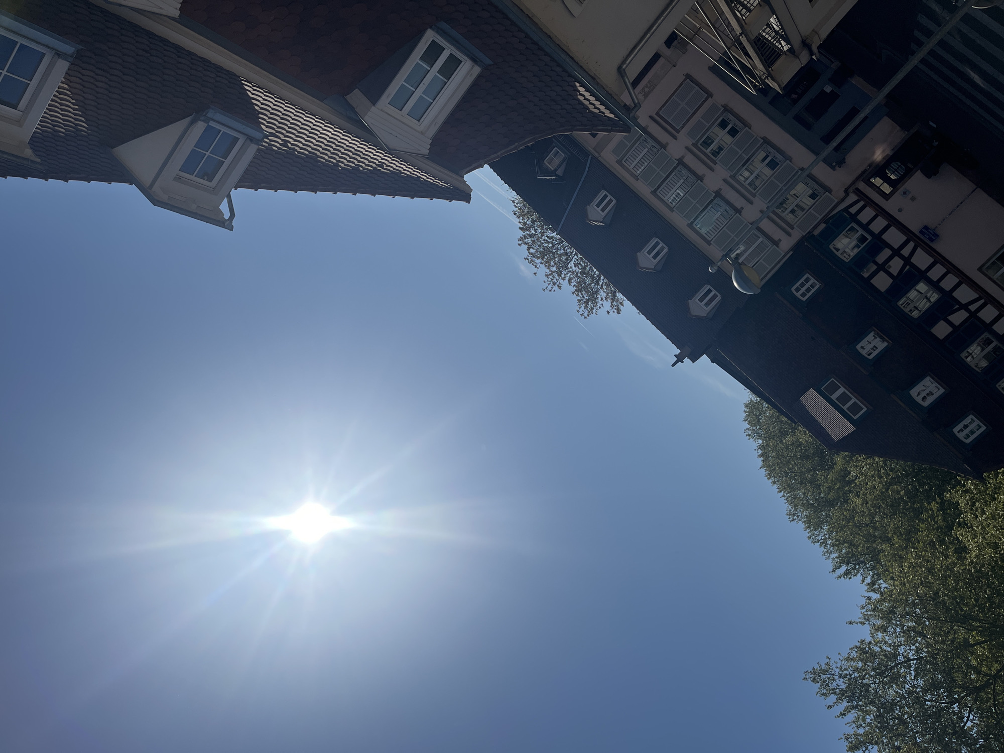 The image presents an upward-looking perspective of buildings and a bright, clear sky in Strasbourg, France. The sun, positioned towards the upper-left when viewed correctly, shines intensely with prominent lens flares and starburst effects, indicating a direct shot into its light. The sky is a vibrant, cloudless blue, suggesting good weather during daytime.
On the left side of the frame, the steep, dark tiled roof of a building is visible, featuring several white-framed dormer windows. One dormer has a distinctive shape with a small gutter. On the right side, a cluster of buildings with varying architectural styles is present. One building has a dark roof with multiple smaller dormer windows, while another features multiple floors with numerous windows, many equipped with white louvered shutters. A traditional half-timbered building, characterized by dark wooden beams against a light-colored facade, is also visible further to the right. A street lamp extends from one of the buildings on the right. Lush green foliage from trees can be seen peeking into the frame from the lower right and between some of the buildings. No human activity is visible, and no legible text can be discerned on the buildings or signs. The architecture is consistent with the historic urban landscape of Strasbourg.