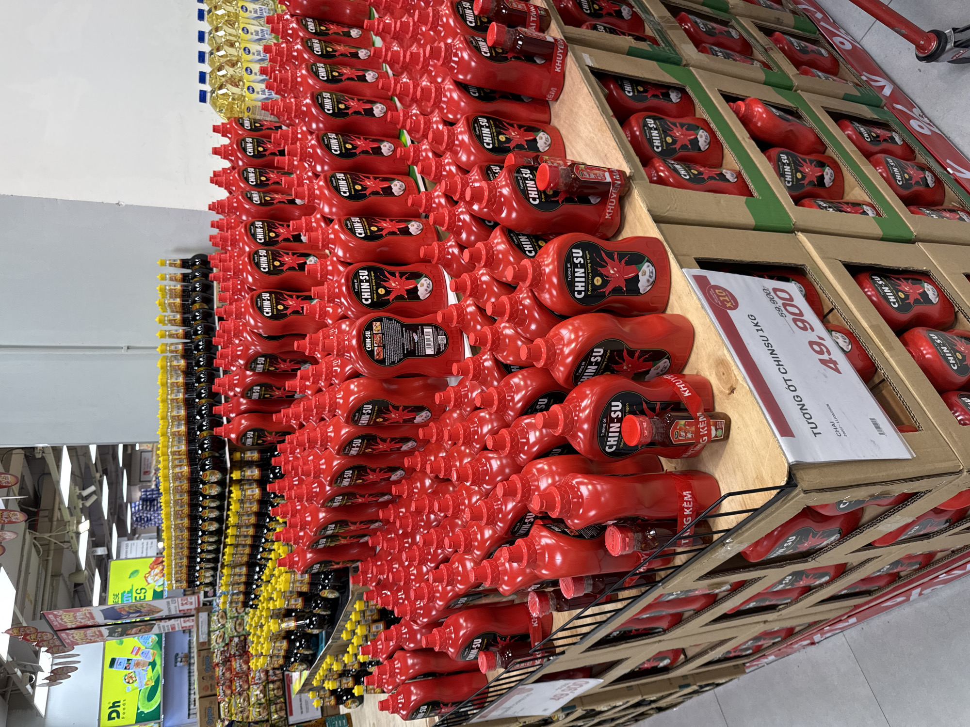 The image depicts a well-stocked aisle within a supermarket in Nha Trang, Vietnam. The primary focus is on a large display of "CHIN-SU" branded products, predominantly chili sauce in vibrant red bottles. These bottles are arranged in multiple tiers on wooden shelves and within cardboard displays, showcasing a significant volume of inventory.
The background reveals more supermarket shelving with various products, including rows of dark bottles with yellow caps, likely soy sauce or similar condiments, and bottles of cooking oil. Fluorescent lighting illuminates the scene from above, reflecting off the glossy surfaces of the product bottles.
Several price tags are visible, including one with the price "49,900" and text indicating "CHINSU 1KG", suggesting a sale or special offer. Another smaller tag shows "X10", possibly indicating a multi-buy discount.
There are no people clearly visible in the immediate foreground or engaged in any specific actions. The environment is clean and organized, typical of a retail grocery store. The overall impression is one of abundance and commercial activity within a Vietnamese supermarket.