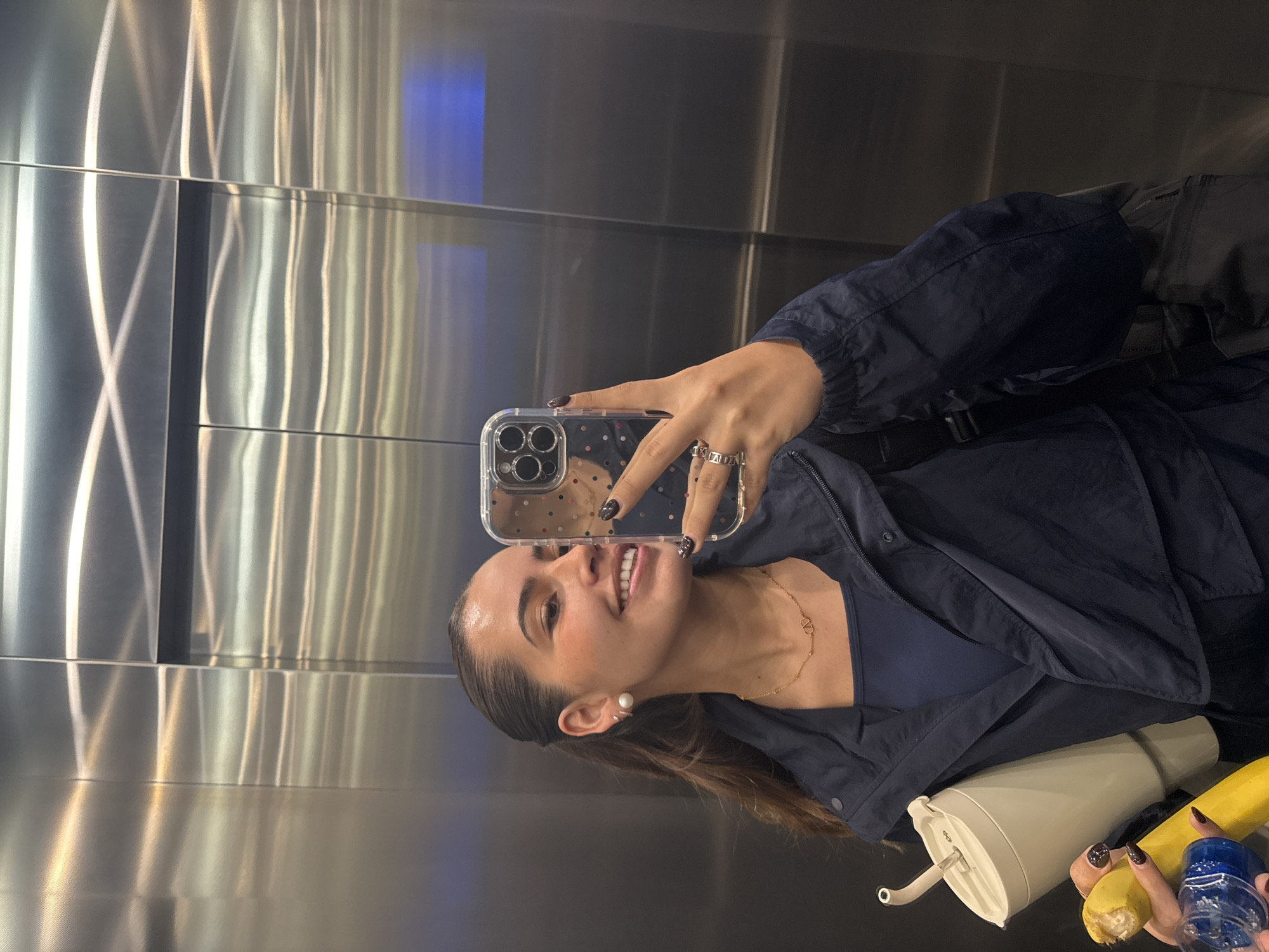 A young woman is depicted in a selfie, taken in what appears to be an elevator or a room with highly reflective, brushed stainless steel walls. The image is rotated 90 degrees counter-clockwise from its original orientation. She is smiling broadly, revealing her teeth, and her expression conveys happiness. Her dark hair is neatly pulled back from her face, with some strands resting on her shoulder.
She is wearing a dark blue jacket over a matching top. Her accessories include pearl stud earrings, an ear cuff, and a delicate gold necklace with a "V" shaped pendant, possibly a designer logo. Her nails are painted a dark, glossy color, perhaps black or deep purple, with a hint of glitter. She holds a smartphone, visible through its clear case adorned with small, colorful polka dots (red, pink, blue, white, black). The phone's triple camera lens setup is clearly visible, reflecting part of her face.
In the lower right corner of the frame, several items are visible in her other hand: a light-colored (beige or cream) tumbler or water bottle with a spout, a yellow banana, and a small blue plastic bottle or container. The metallic walls behind her reflect various vertical light streaks and a distinct blue rectangular light source, contributing to the modern and possibly enclosed setting. While the context mentions Puebla, Mexico, there are no specific visual cues within the image to confirm this location.