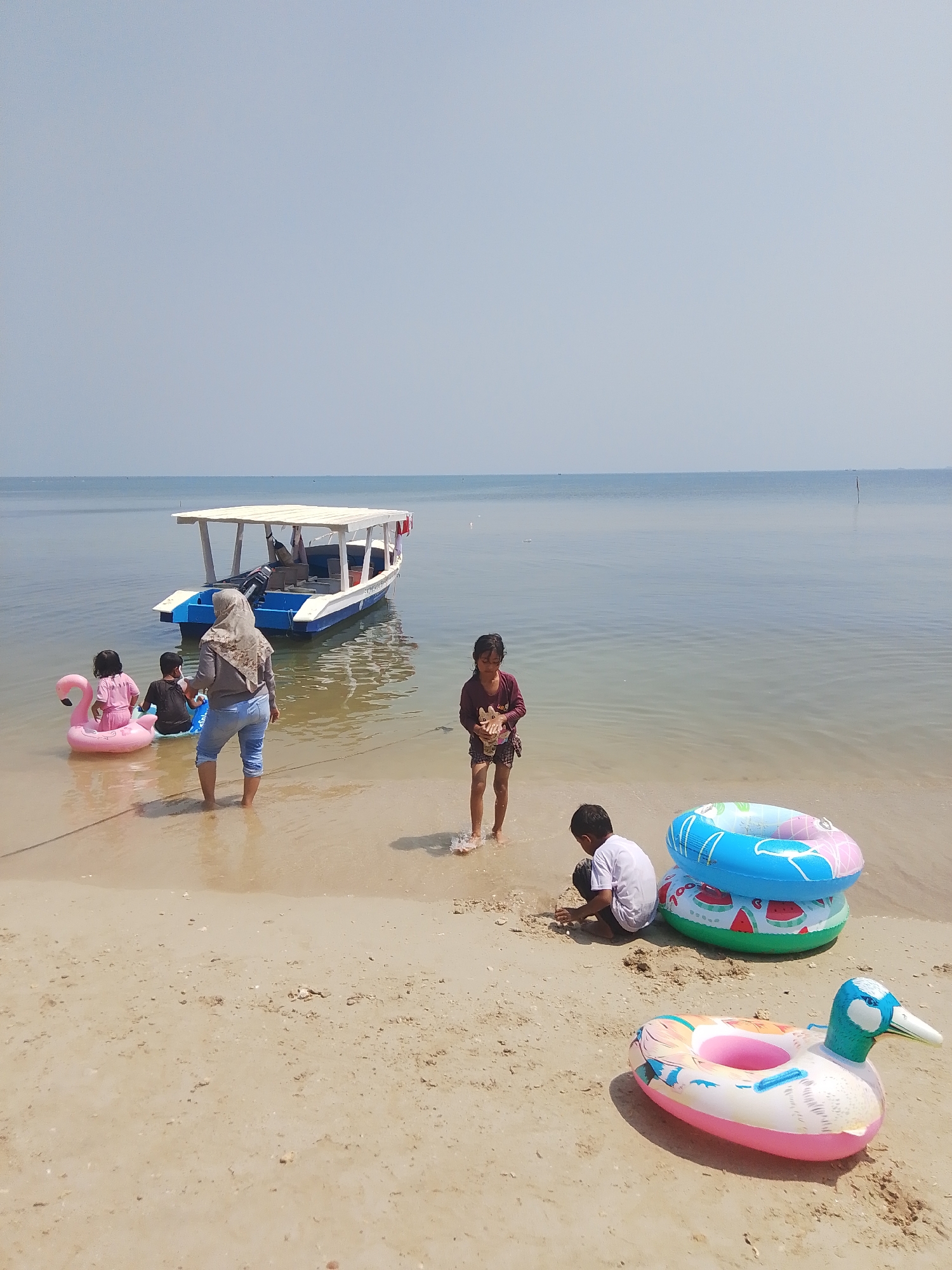In the city of Pejagan, Indonesia, a family enjoys a day at the beach. A white and blue boat is anchored in the calm, shallow water, while several children play on the sandy shore and in the water. One child sits on a pink flamingo float, while another is partially submerged in the water. A woman in a grey hijab and blue jeans stands by the water's edge, facing the children. Two more children are in the foreground, one building a sandcastle and the other sitting near a stack of colorful inflatable rings. A duck-shaped inflatable ring lies on the sand in front of them. The sky is a clear, pale blue, and the horizon is a clean line where the sky meets the sea.