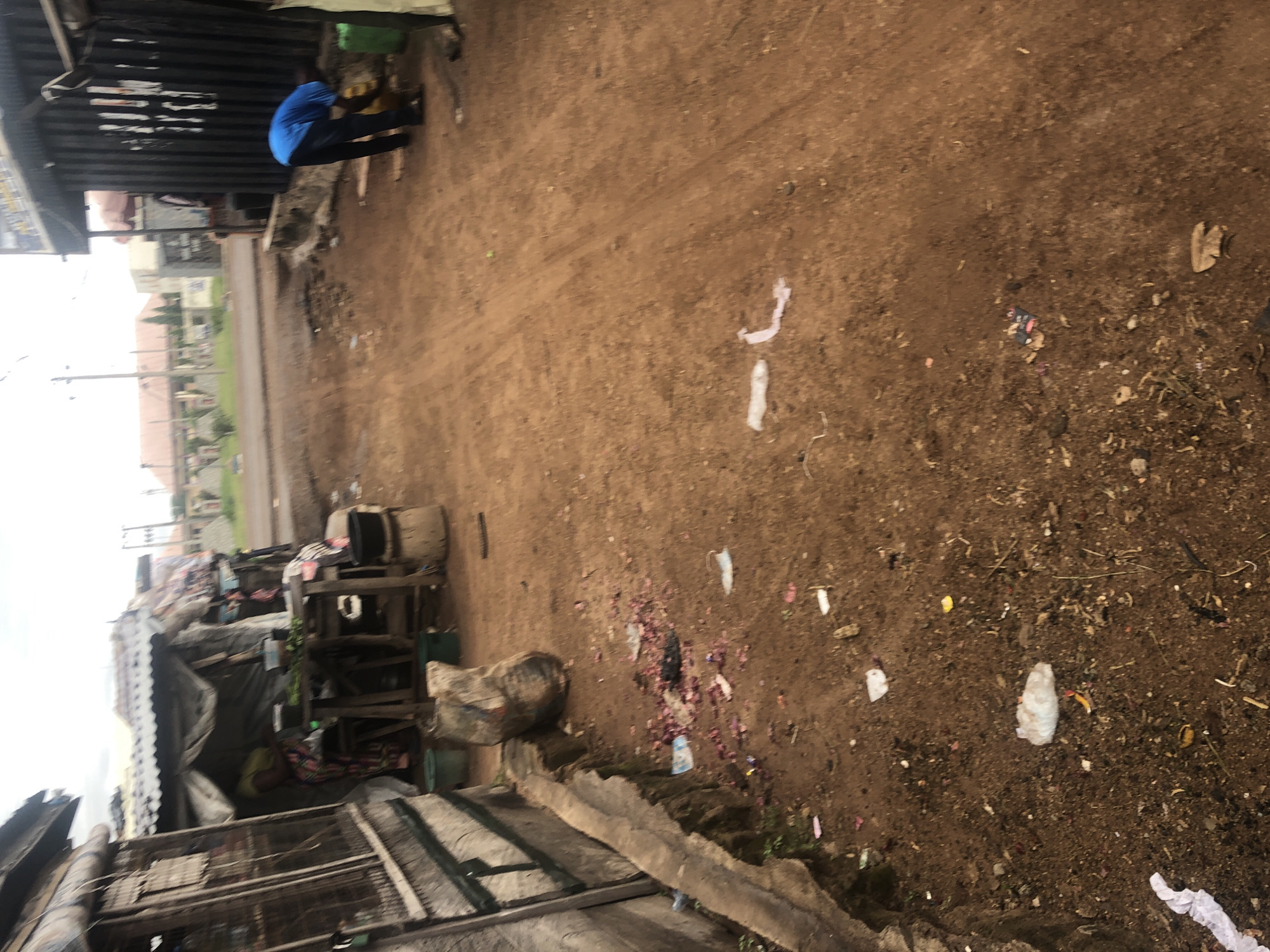The image depicts a dusty, unpaved street in Wukari, Nigeria, likely a commercial or residential area with makeshift structures. On the left, a stall constructed with corrugated metal and wood displays merchandise, with a person standing behind a counter inside. To the right of the stall, a person wearing a blue shirt and dark pants bends down, interacting with a yellow jerrycan, with a green container nearby. The ground is littered with various debris, including plastic bags, wrappers, and what appears to be discarded food items or plant matter. In the background, buildings and power lines suggest an urban setting. The overall impression is one of a working-class area with a street-side economy and potential sanitation challenges.