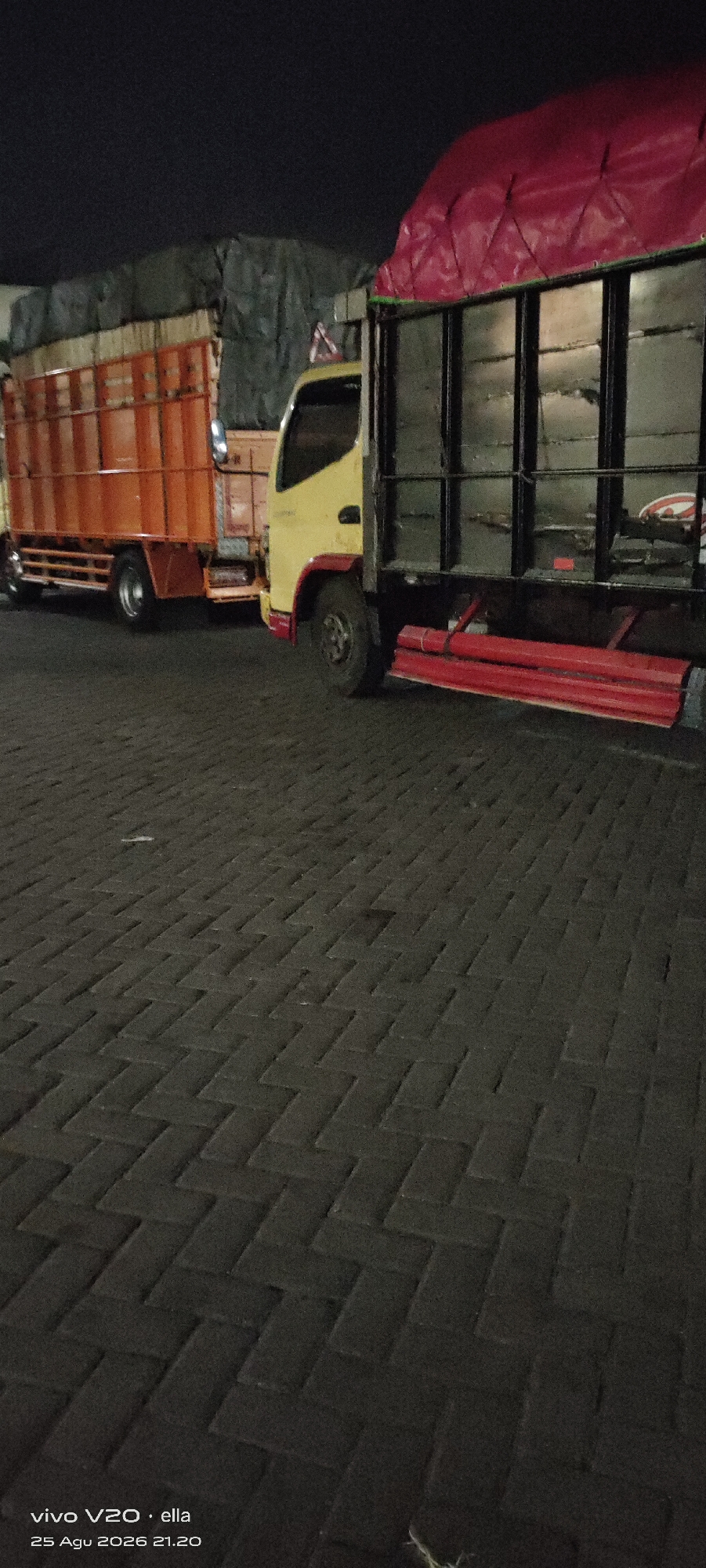 The image shows two trucks parked on a brick-paved surface at night, with a dark sky above. The truck on the left is orange and has a covered cargo area. The truck on the right is yellow and black, with a red tarp covering its cargo. The area appears to be an outdoor space, possibly a parking lot or loading dock. The timestamp at the bottom left of the image indicates it was taken on August 25, 2026, at 21:20, and the location is specified as Tanjungsari, Indonesia. There are no people or visible actions taking place in the scene.