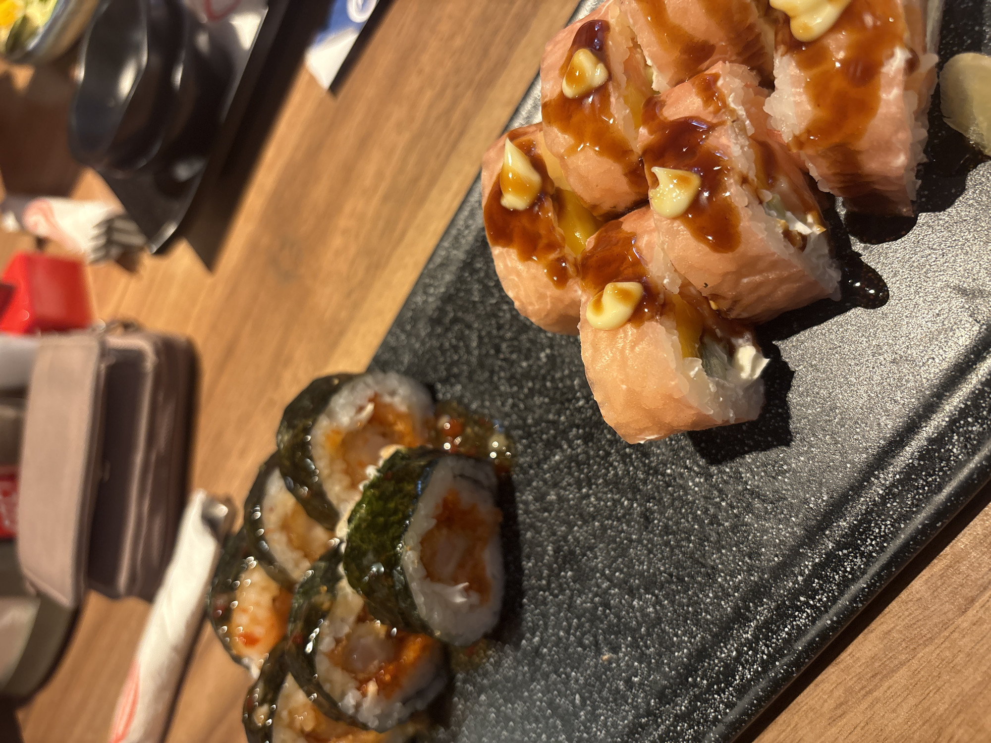 A close-up shot focuses on two plates of sushi on a wooden table. The plate in the foreground, a textured black rectangular dish, holds several pieces of sushi rolls topped with a sauce and a creamy white dollop. To the left, a black square plate holds another arrangement of sushi rolls, partially obscured by the foreground plate. In the background, a light-colored wallet rests on the wooden table, and behind it, out of focus, are other table elements, including what appears to be a red container and a pair of chopsticks. The lighting is warm, casting shadows and highlighting the glistening sauces on the sushi.