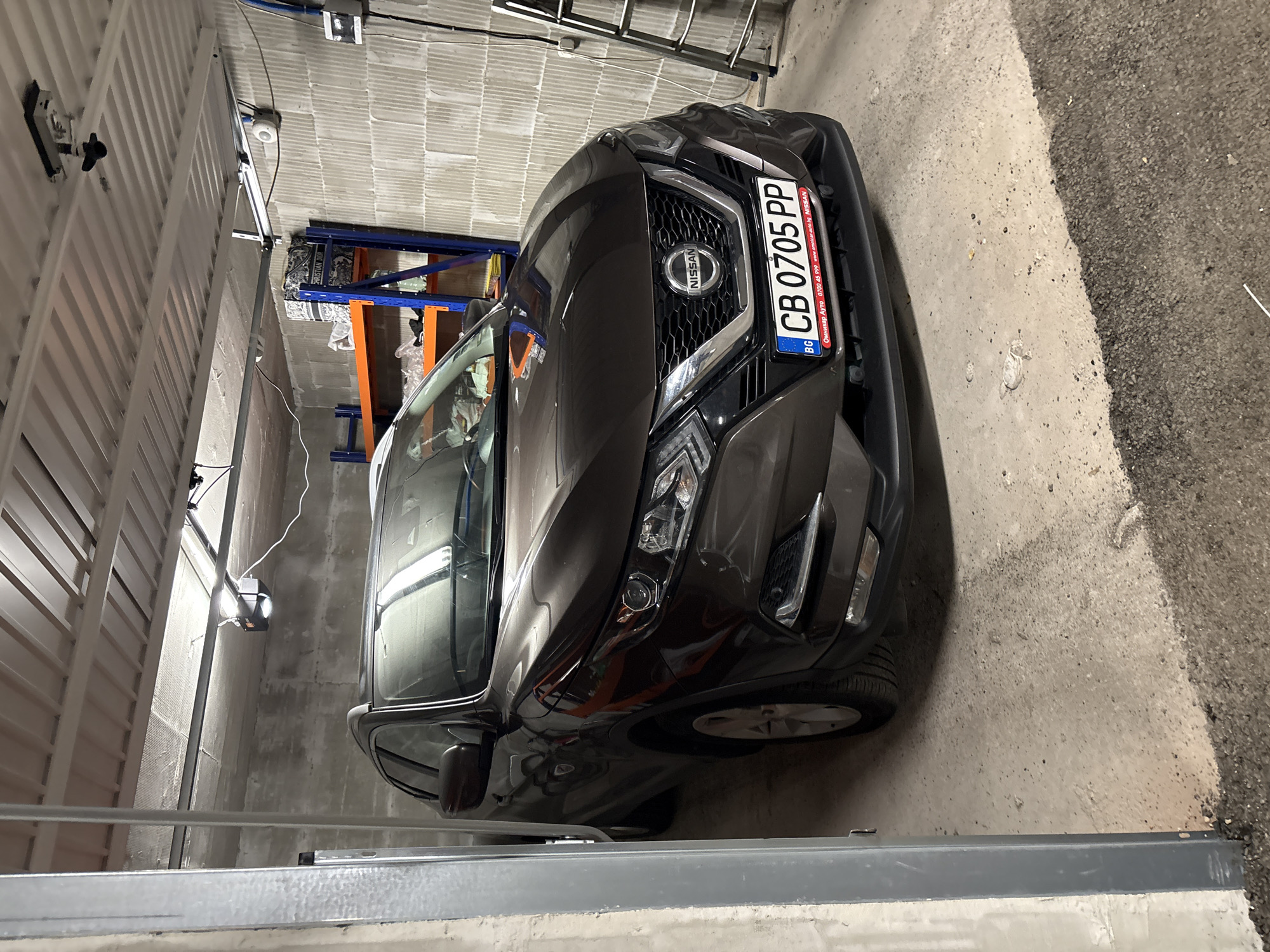 The image depicts a dark gray Nissan Qashqai car parked in what appears to be a garage in Sofia, Bulgaria, as indicated by the license plate "CB 0705 PP". The garage has concrete walls and floor. Storage shelves are visible in the background, along with a garage door opener. The lighting is artificial and appears to be a combination of overhead and natural light. The car is oriented at an angle in the frame.