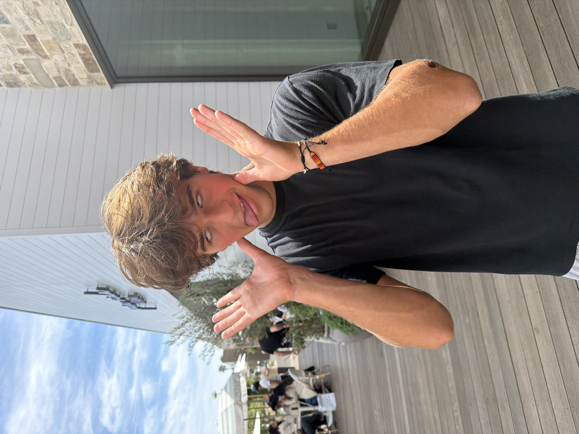 A young man stands outdoors on a wooden deck, making a face with his tongue out and hands held up, fingers splayed. He has light brown hair and is wearing a black t-shirt. On his left wrist, he wears a beaded bracelet. In the background, there are tables and chairs, some with people seated beneath white sun umbrellas. The sky is visible above the scene, a light blue with scattered clouds. The environment appears to be a patio or outdoor dining area, likely located in Malibu, United States of America. The angle of the building's facade, visible on the left side of the frame, further contextualizes the location.