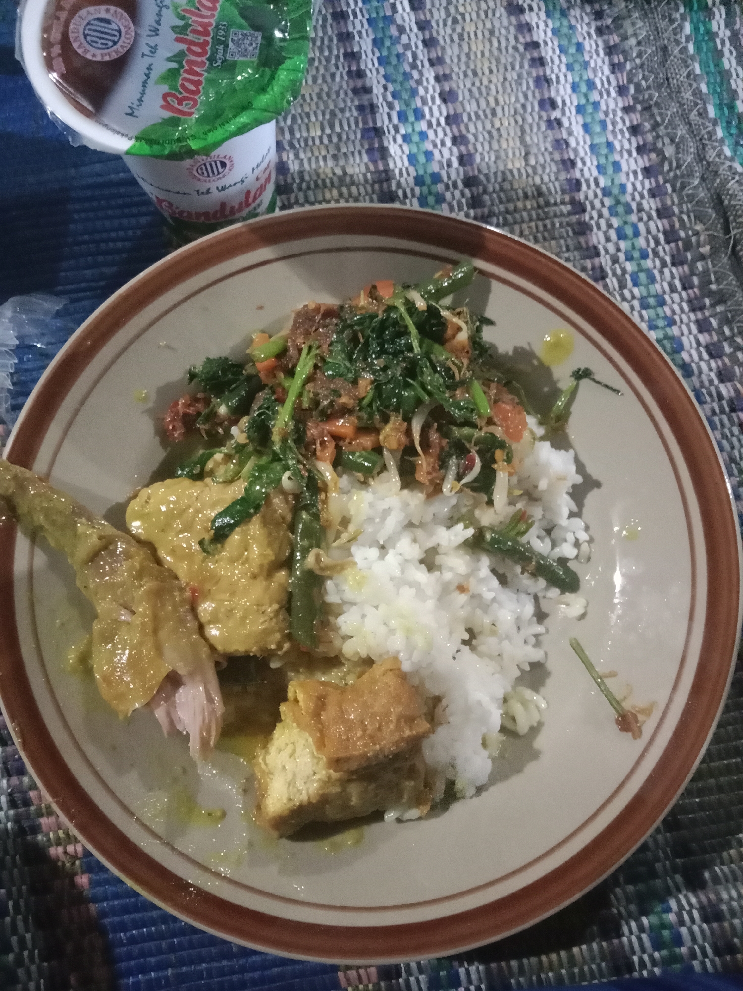 A close-up, overhead shot shows a plate of food and a drink on a woven mat. The plate contains white rice, a piece of yellow-sauced chicken, fried tofu, and a stir-fry of green vegetables and bean sprouts. To the upper left, a cup with a green and white label advertises "Bandulan Minuman Teh Wangi Sejak 1933". The mat appears to be made of blue, white, and grey woven fabric. The scene is likely from a local eatery or home in Brangsong, Indonesia, given the food and branding.