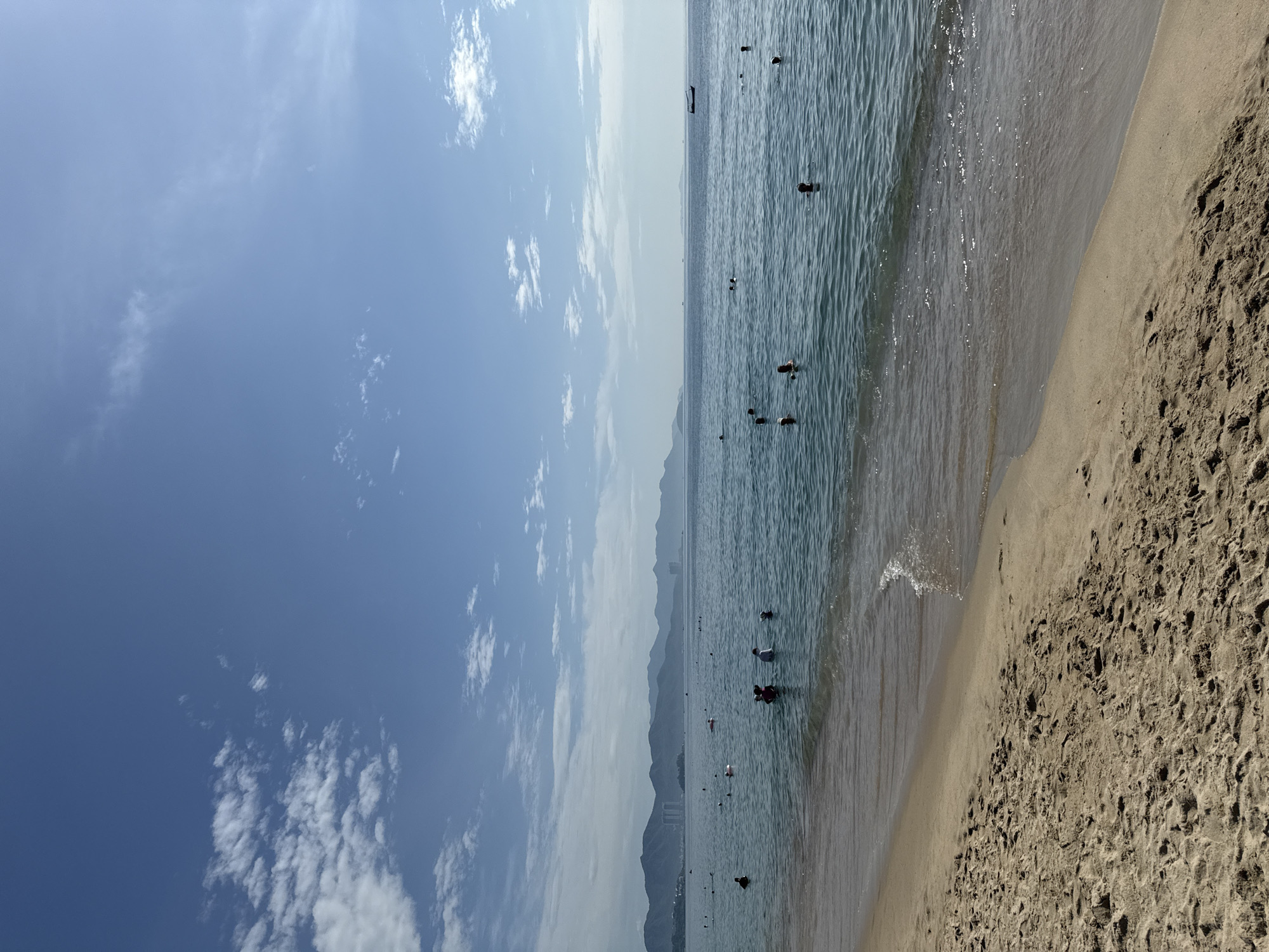The image depicts a panoramic view of a sandy beach in Nha Trang, Vietnam, with the tranquil blue ocean stretching towards a distant, hazy coastline characterized by rolling mountains and scattered buildings. The sky is a vibrant blue, dotted with scattered white clouds, with the sun casting a gentle shimmer on the water's surface. The foreground features a stretch of tan-colored sand, marked with footprints, indicating recent human activity. The ocean is populated by several individuals, seemingly enjoying a swim or wading in the shallow waters. Some are partially submerged, while others are more visible, with their heads and shoulders above the water. A small boat is visible in the distance on the right side of the frame. The overall scene conveys a sense of peacefulness and leisure, typical of a tropical beach destination.