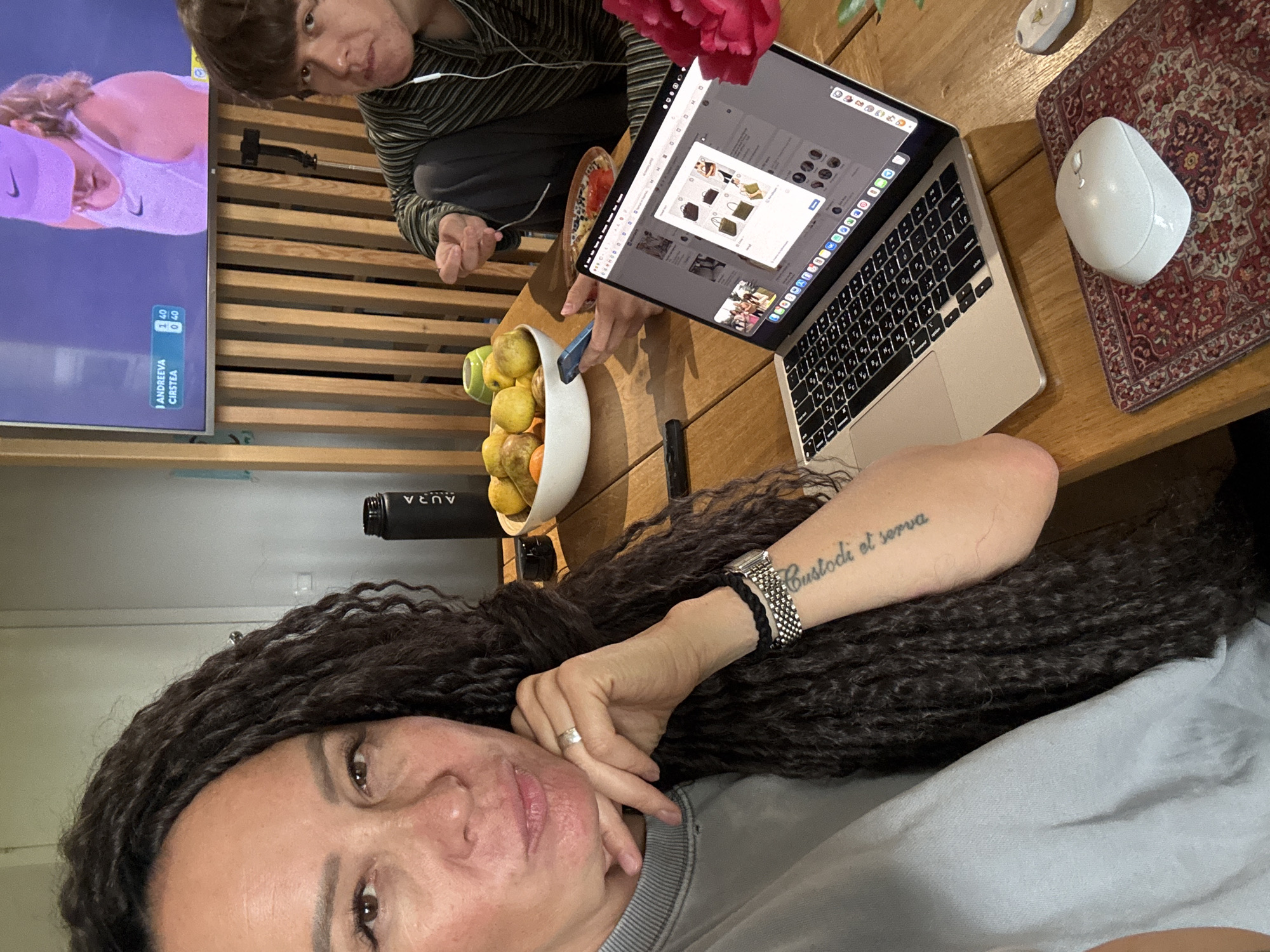 This indoor selfie captures a domestic scene in Chaikovsky, Russia. In the foreground, a person with long, dark, curly hair and a grey top is taking the photo, looking towards the camera with a slight smile. A silver ring is visible on their left hand, and a watch and braided bracelet adorn their left wrist, below a tattoo reading "Custodi et serva."
Behind them, seated at a wooden table, another person with short brown hair, wearing a striped top and wired headphones, looks directly at the camera with a neutral expression. They appear to be resting their arm on the table, near a small plate holding food and a vibrant red flower.
On the table, a silver laptop with a Cyrillic keyboard is open, displaying a webpage, possibly an e-commerce site, showing images of various bags. To the right of the laptop, a white wireless mouse rests on a patterned red and white mousepad. A white bowl filled with oranges and apples sits on the table, and a black bottle or thermos with "AURA" printed on it is also present.
In the background, a flat-screen television is mounted on a slatted wooden wall. The TV is displaying a tennis match, with a female player wearing a white top and a Nike visor visible. The scoreboard shows "ANDREEVA" and "CIRSTEA" with a score of "1 40 | 0 40." The overall setting suggests a relaxed moment at home.