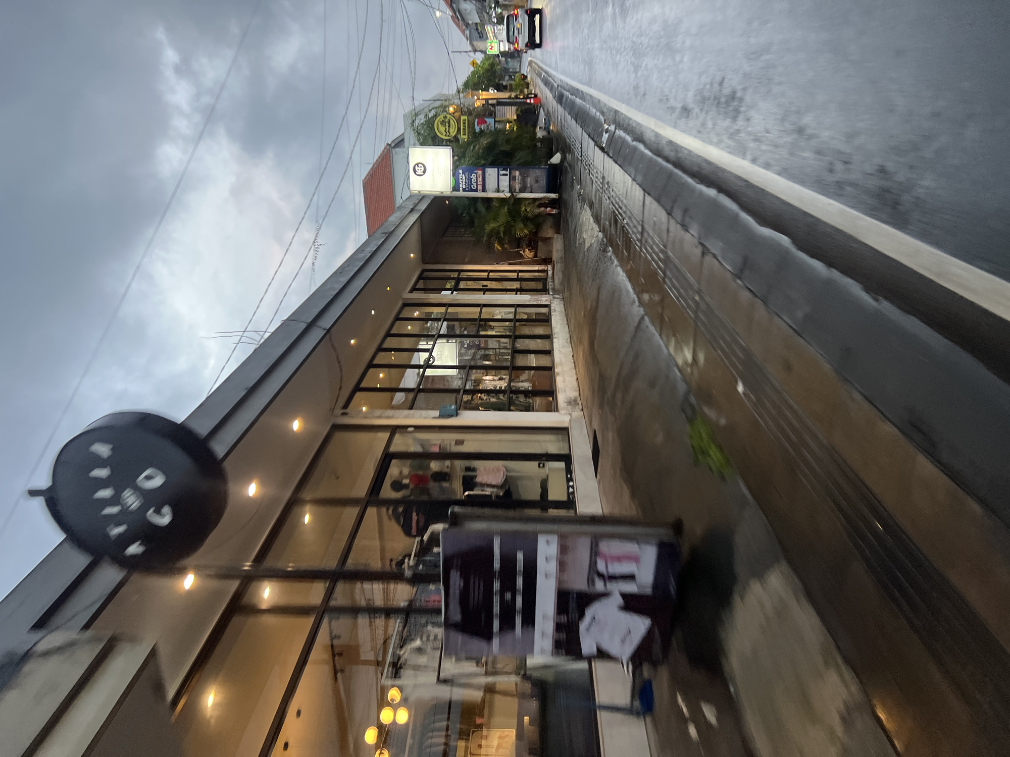 The image depicts a street scene in North Kuta, Indonesia, likely captured during or after rainfall, given the wet pavement. On the left is a modern building with large glass windows, showcasing merchandise inside. A black circular sign with white lettering, reading "ATILIAN CO," is suspended from the building's facade. In front of the building, a display stand features clothing items, including t-shirts. The sidewalk is paved and has a textured surface, indicating accessibility features. A road runs alongside the sidewalk, with a white car visible in the distance on the right. Other signs and vegetation are present along the street. The sky is overcast, suggesting cloudy weather. The perspective suggests the photo was taken from a moving vehicle or while walking.