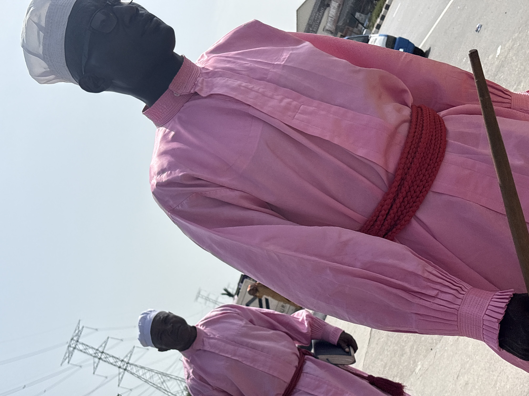 The image captures two figures dressed in pink robes with red rope belts in Igboefon, Nigeria. The foreground figure wears a white cap and holds a brown staff. In the background, another figure, also wearing a white cap and pink robe, carries a book. Overhead power lines and signage are visible in the distance, suggesting an urban environment. The background also shows vehicles on what appears to be a road.