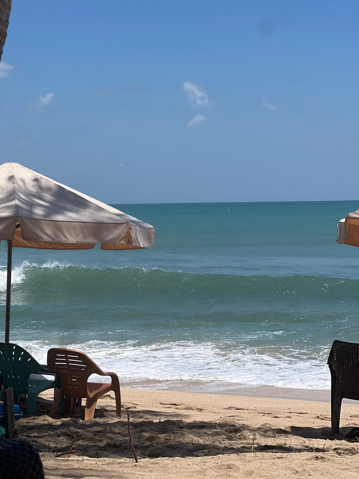 The image depicts a serene beach scene on a clear, sunny day. In the foreground, on a sandy beach, are several beach chairs, some in green and brown, positioned under a light-colored umbrella. The ocean stretches out to the horizon with calm, turquoise water and gentle waves breaking on the shore. The sky is a bright, cloudless blue, with a faint, distant airplane visible. To the right, a portion of another umbrella is visible. The overall atmosphere is one of relaxation and tranquility, characteristic of a popular tourist destination like Kuta, Indonesia.