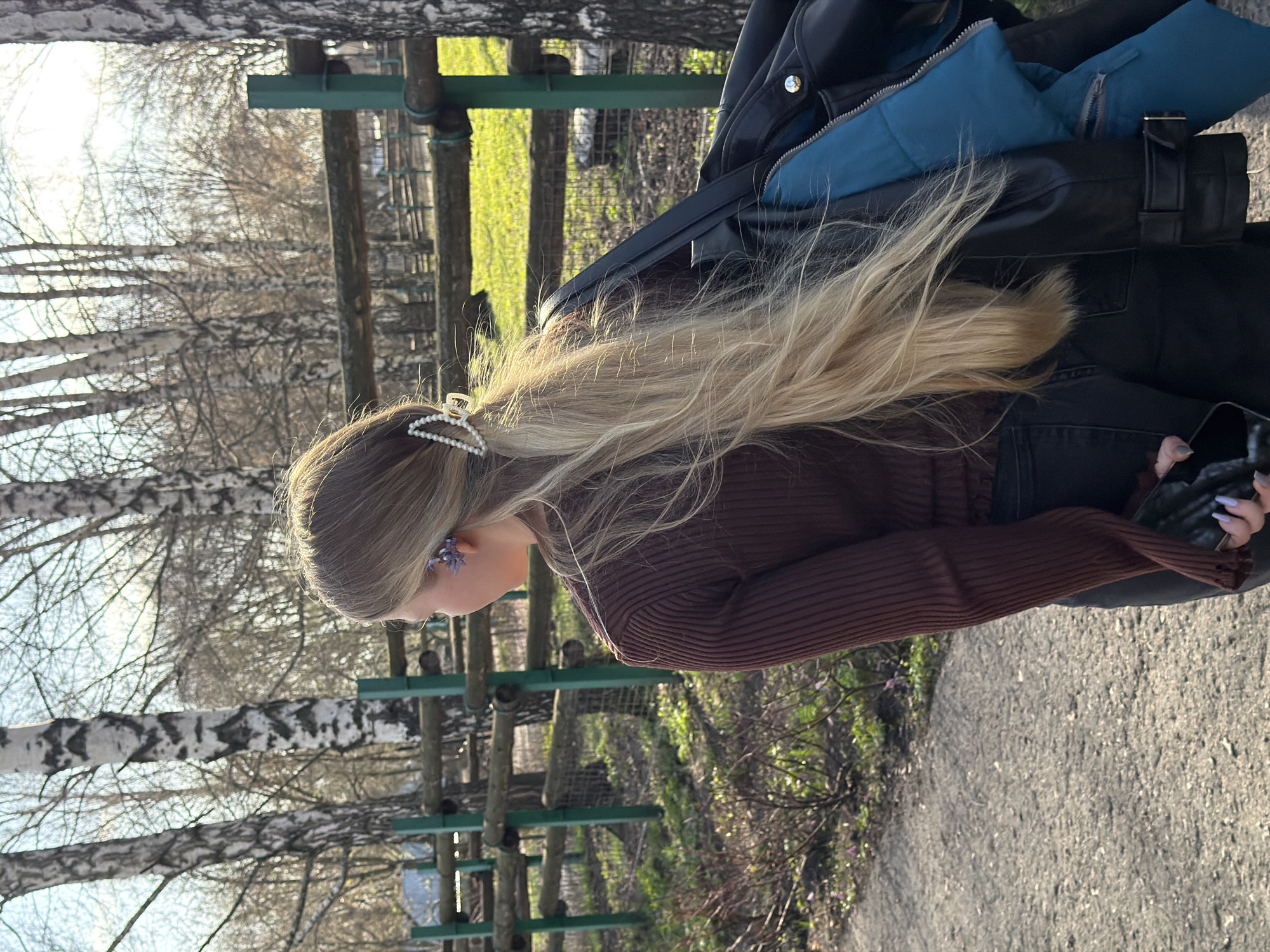 A young woman is seen in a medium shot from the side, outdoors. She has long blonde hair, styled with a pearl hair clip towards the back of her head. She wears a dark brown, long-sleeved, ribbed sweater, dark jeans and carries a black leather jacket. She also has blue floral earrings. In her hand, she holds a mobile phone with light purple nails. The background features a wooden playground or obstacle course structure set amongst bare trees, which are indicative of a late winter or early spring season. The ground is paved, and sparse vegetation is visible. The location is Penza, Russia.