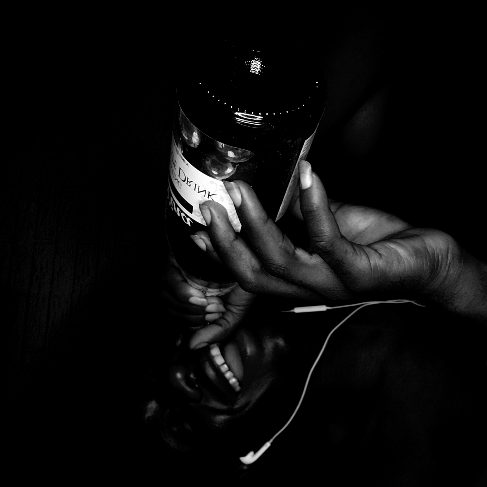 A black and white photograph shows a hand holding a bottle labeled "LE DRINK" above a reflective surface. The reflection shows a smiling face with earbuds in. The environment is very dark. There is no indication that the location is Narayi, Nigeria.
