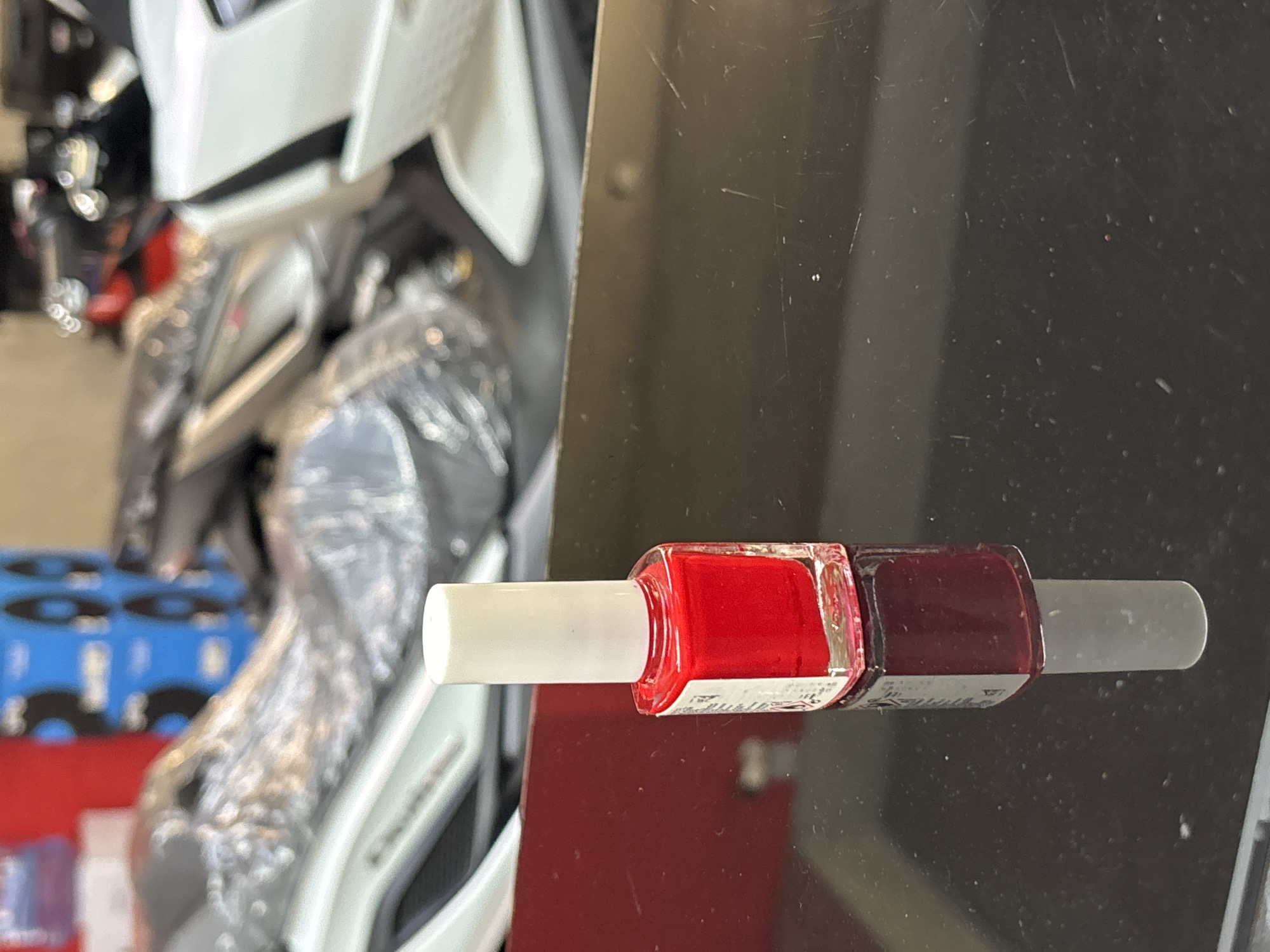 A red nail polish bottle stands on a black reflective surface. The bottle has a white cap and a small white label with some text. The background is blurred and shows what appears to be motorcycles wrapped in plastic. The scene is lit by artificial light. There are no people visible in the image.