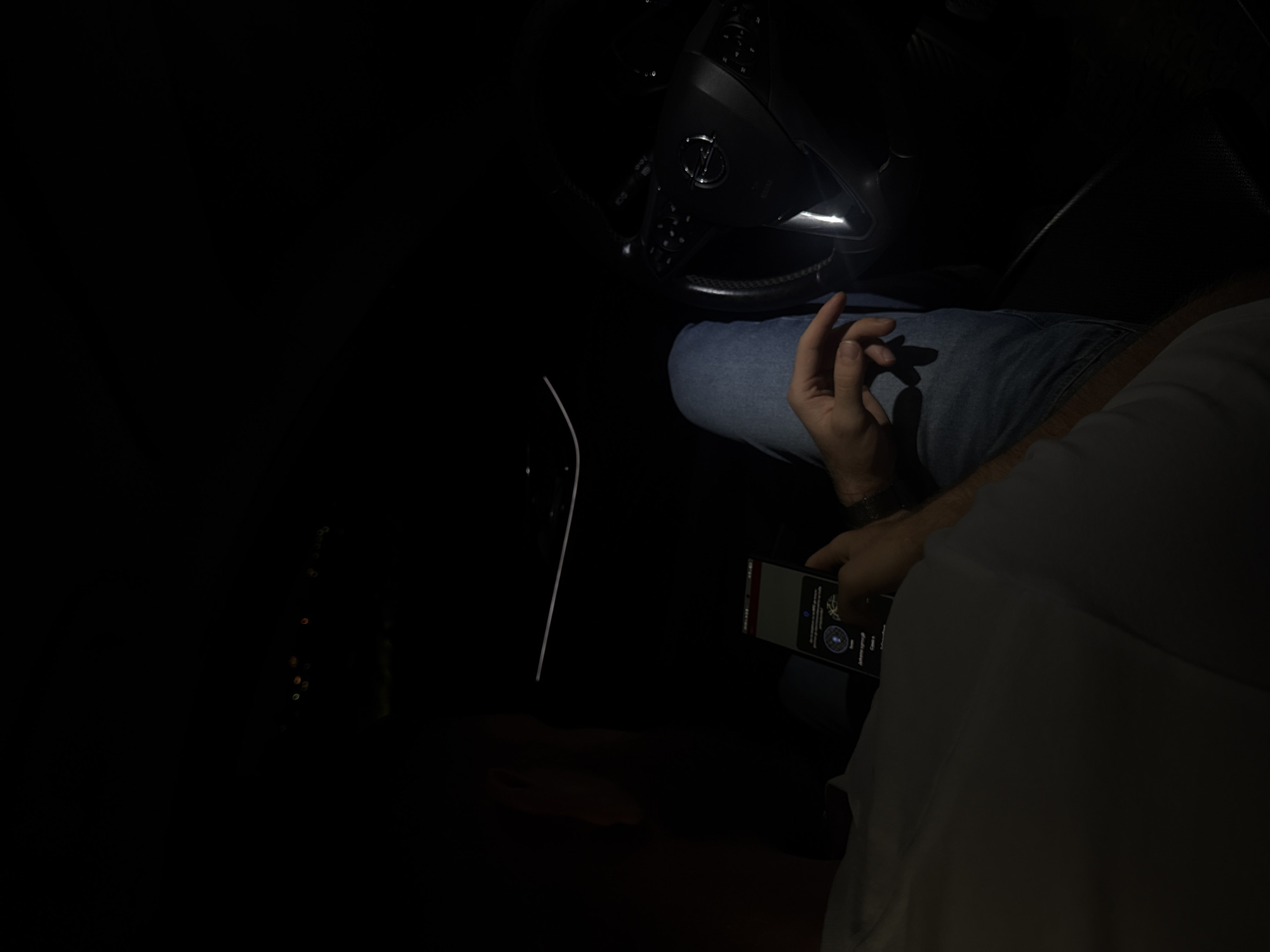 The image captures the interior of a vehicle during nighttime or in an extremely low-light environment. A person, largely obscured by shadow, is visible from the chest down, wearing a light-colored top and blue denim jeans. This individual is seated, likely in the driver's position, and is actively engaging with a black smartphone held in their left hand, with a finger or thumb from their right hand touching the screen. The phone's illuminated display shows text in Cyrillic script, which could be Bulgarian, Serbian, or Macedonian, along with a circular graphical interface element. To the right, the dark steering wheel of a modern Opel vehicle is clearly visible, featuring the brand's logo in the center and multi-function buttons on its spokes. Faint, out-of-focus lights, possibly dashboard indicators or reflections, are discernible in the upper background. The external environment through the windows is completely dark and devoid of any identifiable features. Based solely on the visual information within the image, there are no discernible landmarks, street signs, or unique architectural elements to confirm the specific location as Varna, Bulgaria.