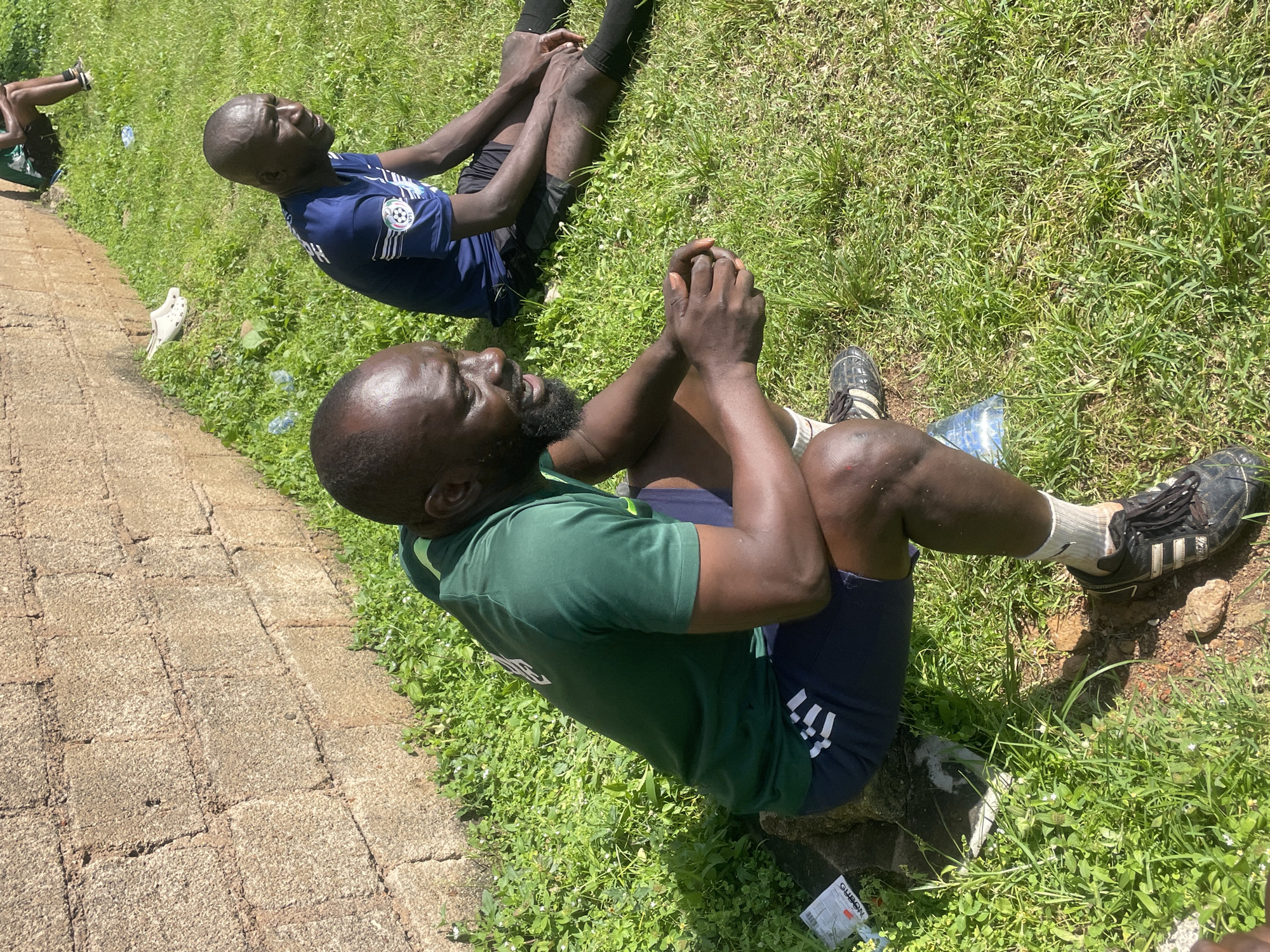 Two men are sitting on a grassy area next to a brick wall in Jos, Nigeria. The man in the foreground is wearing a green t-shirt and dark shorts. He has a beard and is looking to the side with his mouth open as if speaking. The man in the background is wearing a blue t-shirt and black shorts. He is looking to the side with his mouth closed. There are several plastic water bottles scattered on the grass. The sun is shining and casting shadows.