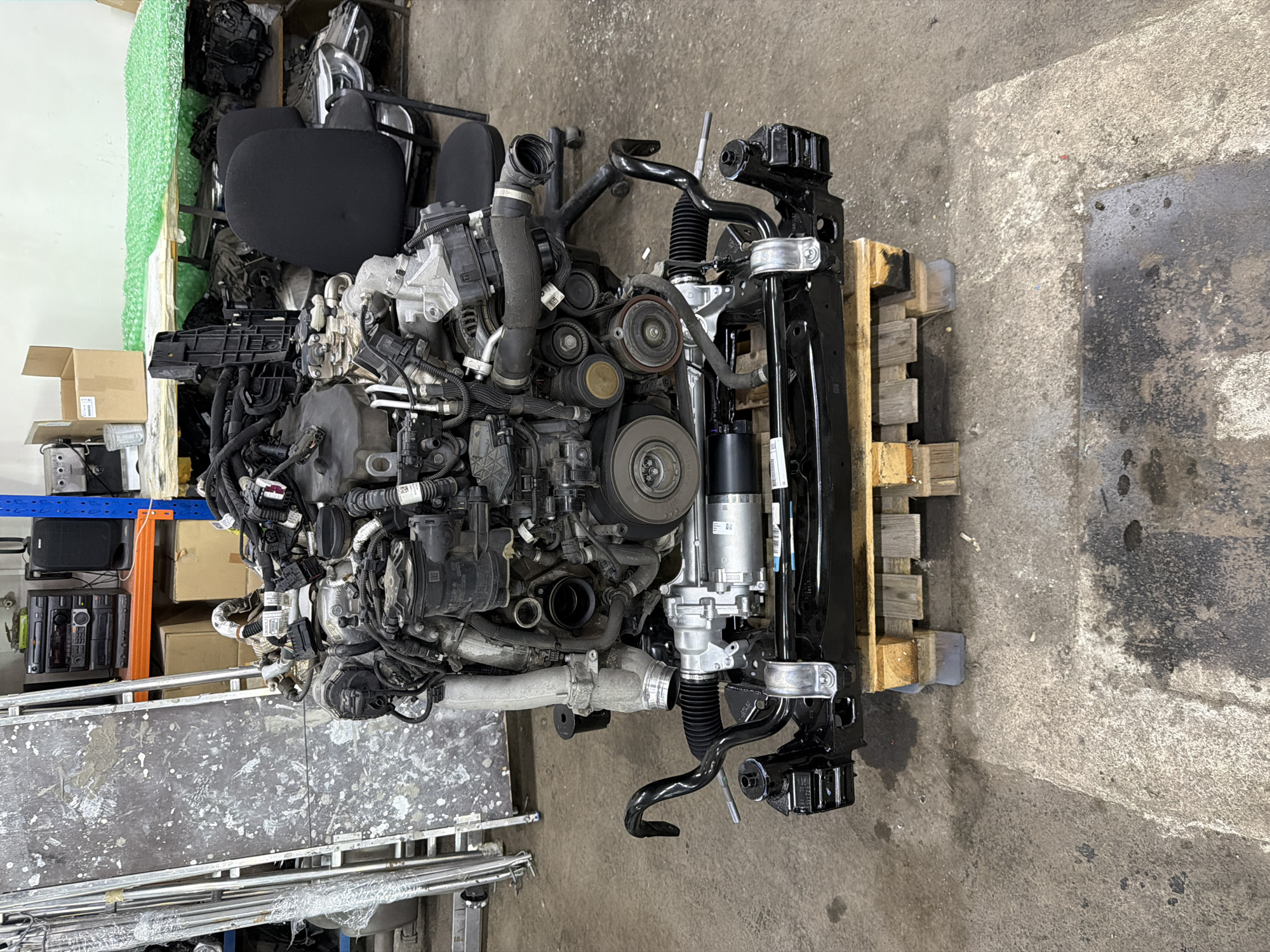 The image shows an engine, possibly a car engine, mounted on a wooden pallet. The engine is a complex arrangement of metal components, including belts, hoses, and wiring. It appears to be used or removed from a vehicle. The pallet is situated on a concrete floor. Other objects in the background include storage shelves with various items like car parts, boxes, and equipment. The location is likely a garage or automotive repair shop. I do not see anything in the image that can confirm that this location is specifically Burgas, Bulgaria.