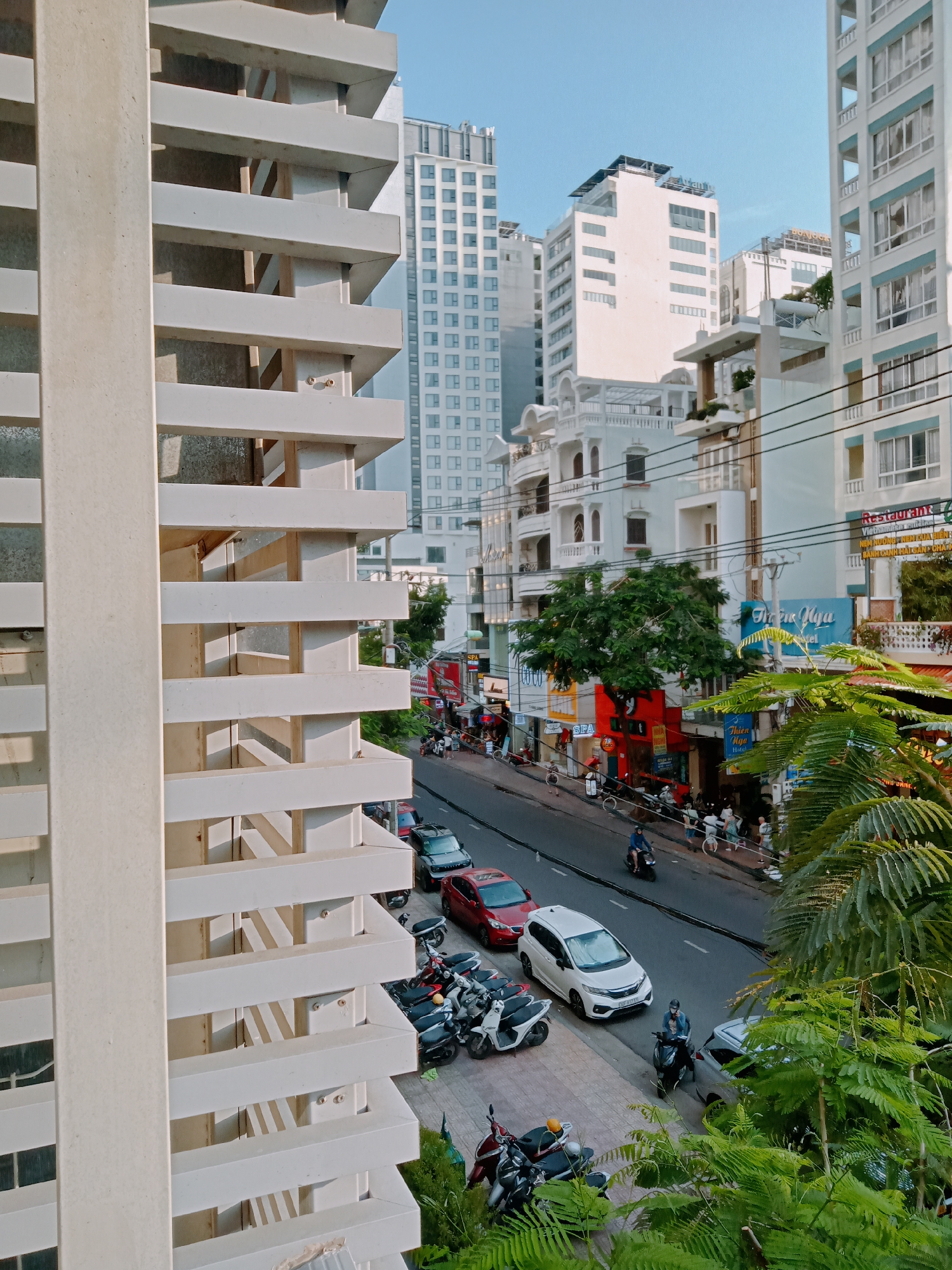 The image shows a street scene in Nha Trang Ward, Vietnam, viewed from an elevated position through a window with horizontal white louvers. The street contains cars, motorcycles, and pedestrians. Buildings line the street, including tall, white structures. The sky is a clear blue. Lush green foliage is visible in the foreground, near the bottom right corner of the frame.