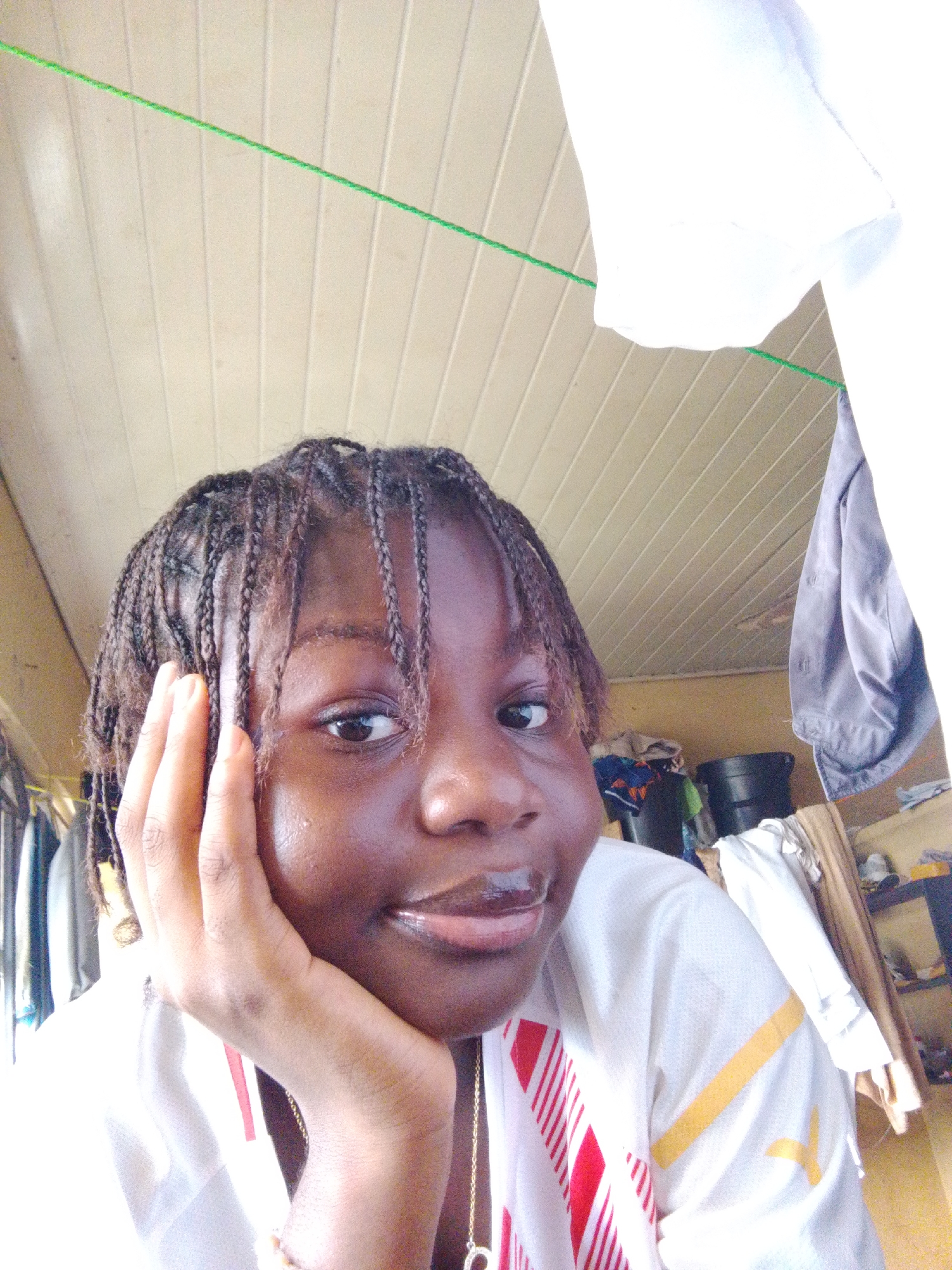 A person with dark skin and braided hair is shown from a low-angle perspective indoors. The person's left hand is resting on their face, and they are wearing a white shirt with red and yellow stripes. A gold chain necklace is visible. Behind the person is a horizontal white ceiling with a green string running across it. In the background, there are clothes hanging on a rack and other items stacked on shelves, suggesting a cluttered room. The location is possibly a private residence in Nsukka, Nigeria.
