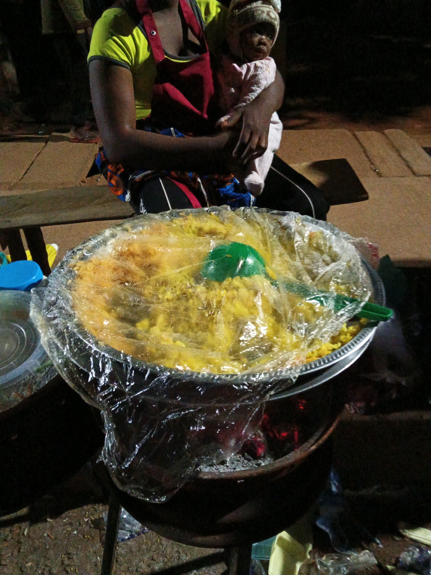 A woman, her arms dark-skinned, is holding a baby wrapped in a white garment and a knitted cap. She is wearing a bright green t-shirt and a red apron over a patterned skirt. In front of her, on a metal stand over coals, is a large, round metal bowl filled with yellow food, possibly jollof rice, covered in clear plastic wrap. A green plastic serving utensil rests on top of the food. The scene appears to be outdoors, possibly a street vendor's stall in Bukuru, Nigeria. The ground is a mix of dirt and possibly paving stones, and there are wooden planks in the background. The lighting suggests it is evening or night.