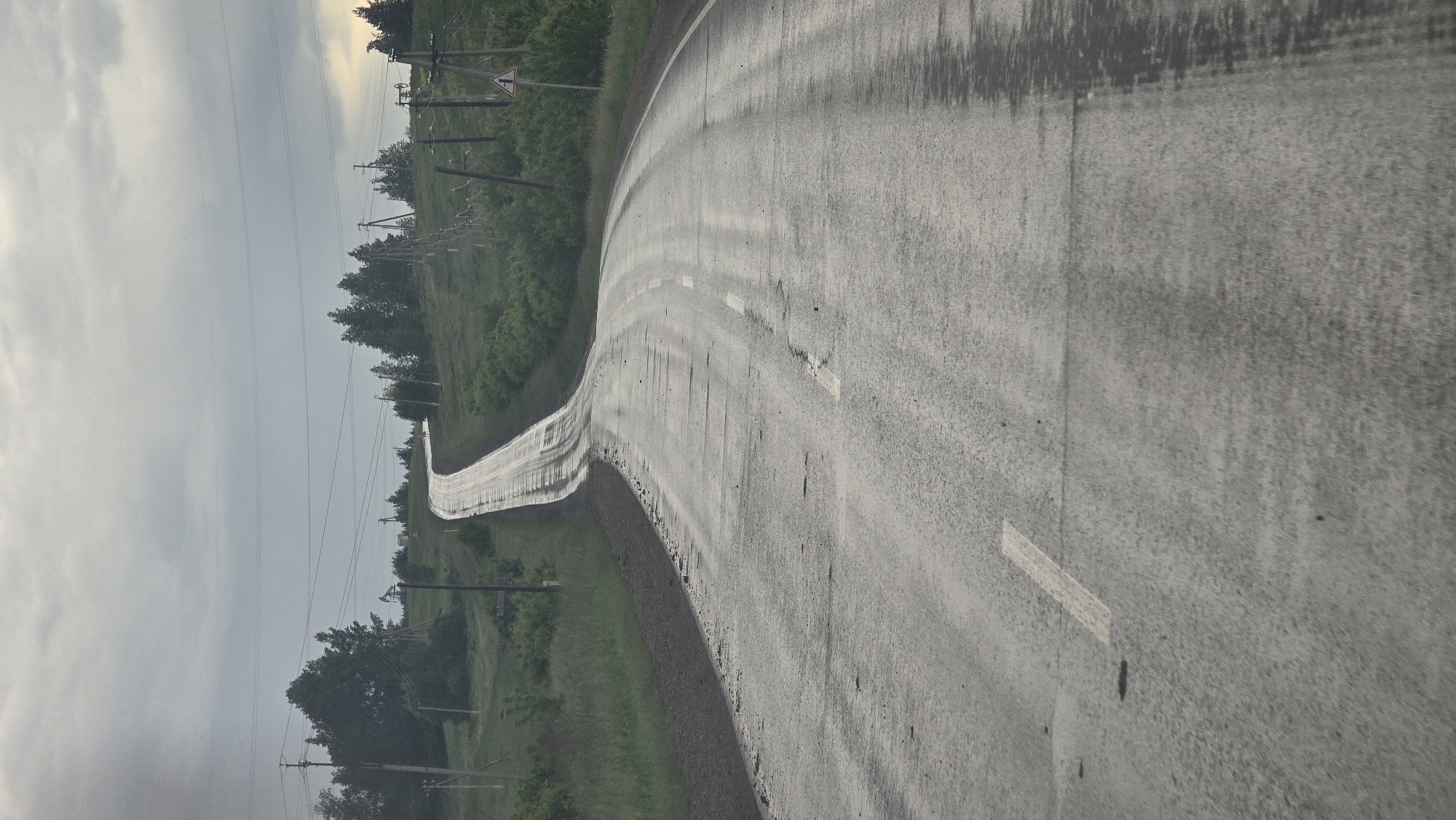 This image depicts a two-lane asphalt road under an overcast sky in Leninogorsk, Russia. The road surface is wet, showing reflective patches, particularly in the distance, and exhibits signs of significant wear, including numerous cracks, unevenness, and potholes. A faded, dashed white line marks the center of the road. The road gently curves to the right in the foreground and mid-ground, then ascends sharply and curves to the left in the distance, disappearing over a crest. The right shoulder of the road in the foreground is dark and slightly elevated. Both sides of the road are flanked by green vegetation, consisting of grass, bushes, and scattered trees, with denser tree clusters appearing further uphill. Numerous utility poles carrying power lines run parallel to the road on both sides. On the right side of the road, in the mid-ground, a white triangular warning sign with a black border and an internal black symbol (likely indicating an uneven road or other hazard) is visible. In the distant left, amidst the trees and utility poles, an oil pumpjack is discernible. No people or vehicles are present in the scene. The lighting is diffused, consistent with an overcast day.