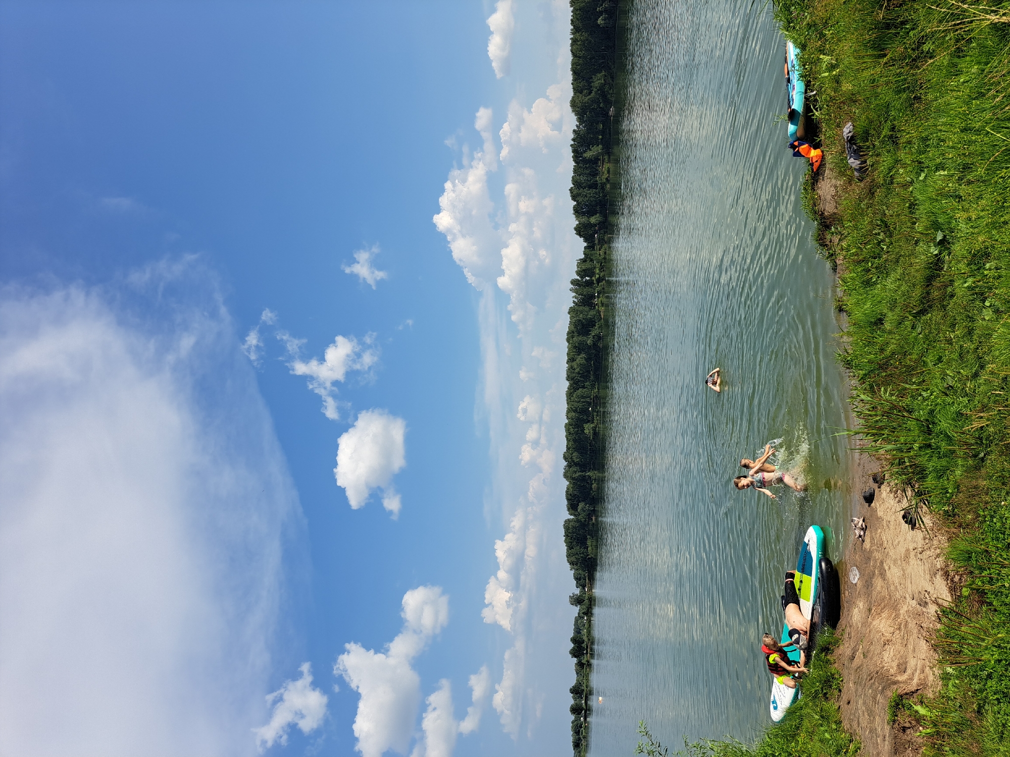 In Sorochii Log, Russia, on a sunny day with scattered white clouds against a blue sky, a group of individuals are engaged in water activities by a lake. In the foreground, on the grassy bank, are two paddleboards. One paddleboard has a person lying on it wearing a life vest and swim trunks, and another person sitting on the edge with a life vest. Near them, a black inflatable ring is partially submerged in the water. Further into the lake, two children are splashing and playing, with one child appearing to jump. Another person is visible in the distance, swimming. On the right side of the bank, a blue kayak with orange life vests is present. The background features a dense line of green trees across the lake, suggesting a natural landscape. The water is calm with ripples.