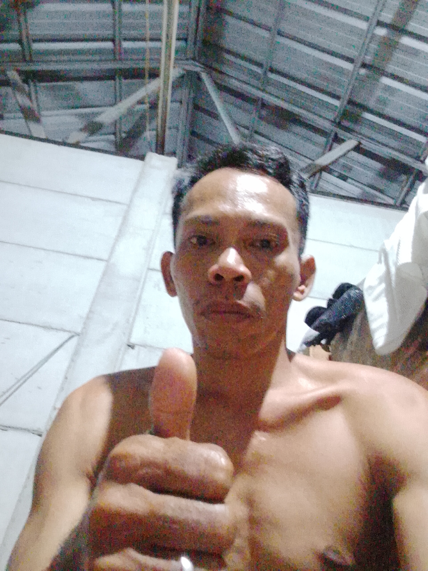 A shirtless man in Bukit Ketok, Indonesia, gives a thumbs-up. He stands indoors, under a ceiling of corrugated metal and exposed beams. White walls are behind him. Some dark and white clothing is in the background.