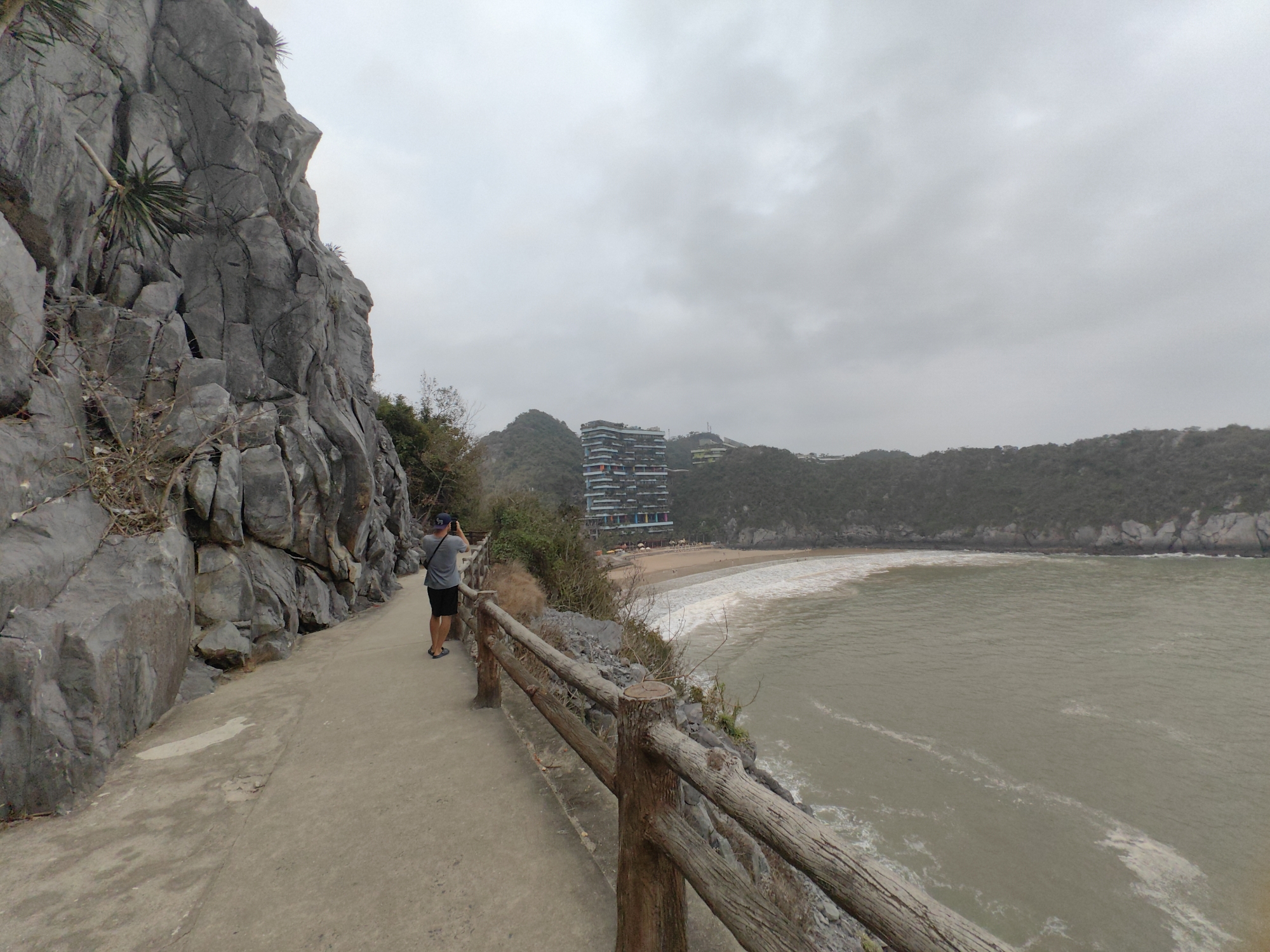 A wide-angle shot depicts a coastal scene on an overcast day in Hải Phòng, Vietnam. A man stands on a concrete walkway, facing the ocean and holding a mobile device to capture a photograph. The walkway is bordered by a weathered wooden railing and a rocky cliff face on the left side. The water of the bay is murky, with visible wave action. In the background, a multi-story building with a unique, colorful facade stands on a hilltop overlooking a sandy beach. Additional forested hills and a cloudy sky complete the composition.