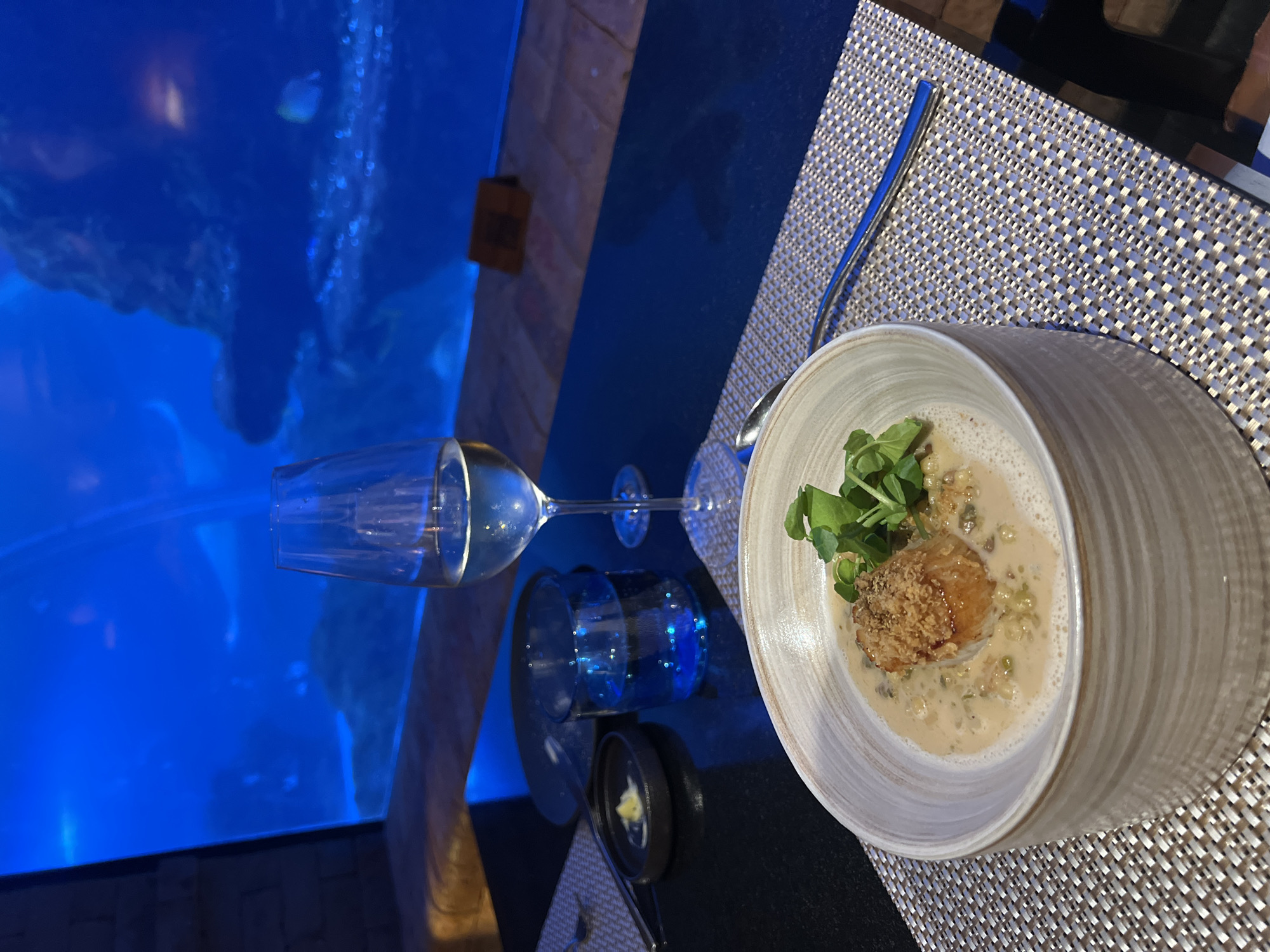 The image captures a dining scene in Kuta Selatan, Indonesia. In the foreground, a bowl of soup is prominently displayed on a woven placemat. The soup appears to contain a fried element, garnished with fresh greens. Next to the bowl is a spoon. A wine glass lies horizontally, its contents reflecting the ambient blue light. In the background, a large aquarium filled with blue water is visible, suggesting the restaurant may feature marine life as part of its ambiance. The scene presents a combination of culinary presentation and environmental atmosphere.