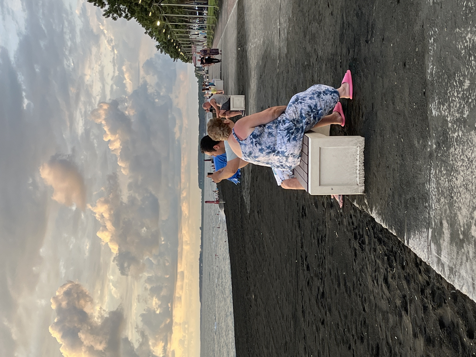 In Ureki, Georgia, a couple is seated on a bench on a black sand beach beside a promenade. The man, wearing a blue t-shirt, is taking a selfie with a smartphone. The woman, dressed in a blue and white floral jumpsuit and pink slippers, is sitting next to him. The sea stretches out behind them, with a few people visible in the water. The promenade, lined with streetlights and trees, runs along the edge of the beach, with other individuals walking and sitting on benches in the distance. The sky is filled with dramatic clouds, illuminated by the setting sun.