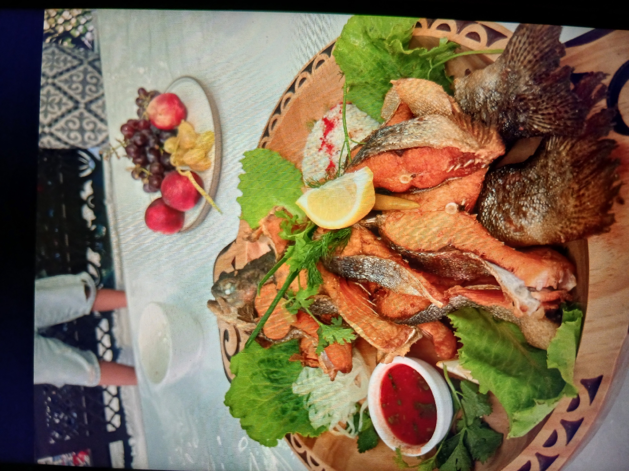 In Yanta District, China, a close-up, top-down shot captures a wooden platter piled high with fried fish, garnished with lettuce leaves, a wedge of lemon, and a small bowl of red sauce. A bunch of grapes and some red apples are visible on a plate in the background, placed on a table covered with a light-colored tablecloth. In the far background, the legs of a person wearing light-colored shorts are visible, with their feet submerged in water, possibly in a shallow pool or a decorative water feature. The scene appears to be outdoors, with a patterned screen or railing behind the person.