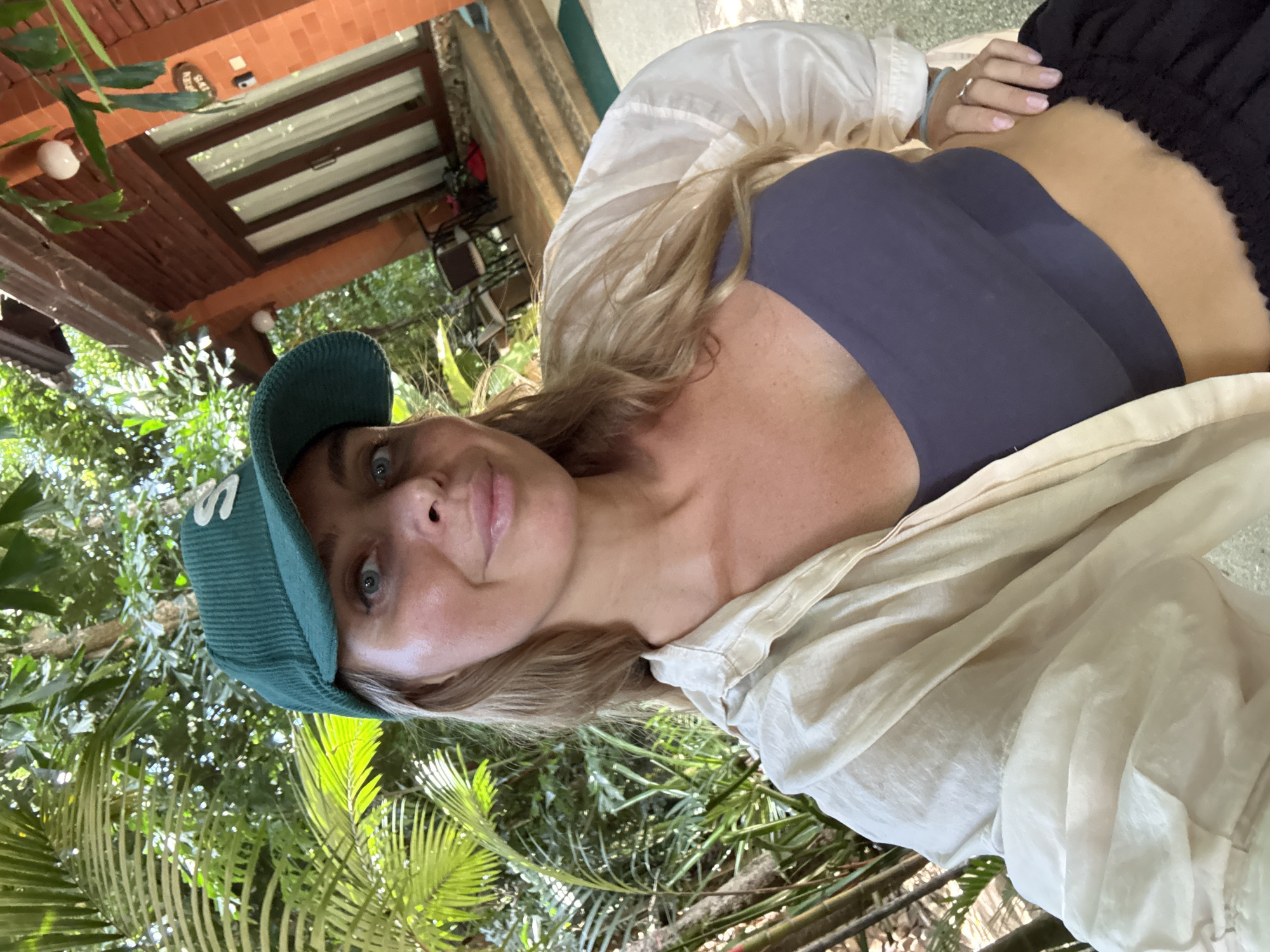 In the city of Ko Samui District, Thailand, a woman with blonde hair and blue eyes is taking a selfie. She is wearing a green corduroy baseball cap with a white 'S' logo, a light-colored button-up shirt worn open, and a dark blue tank top. She is outdoors, surrounded by lush green foliage. In the background, a building with an orange brick facade and a wooden porch is visible, with stairs leading up to a doorway. The scene suggests a tropical vacation setting.
