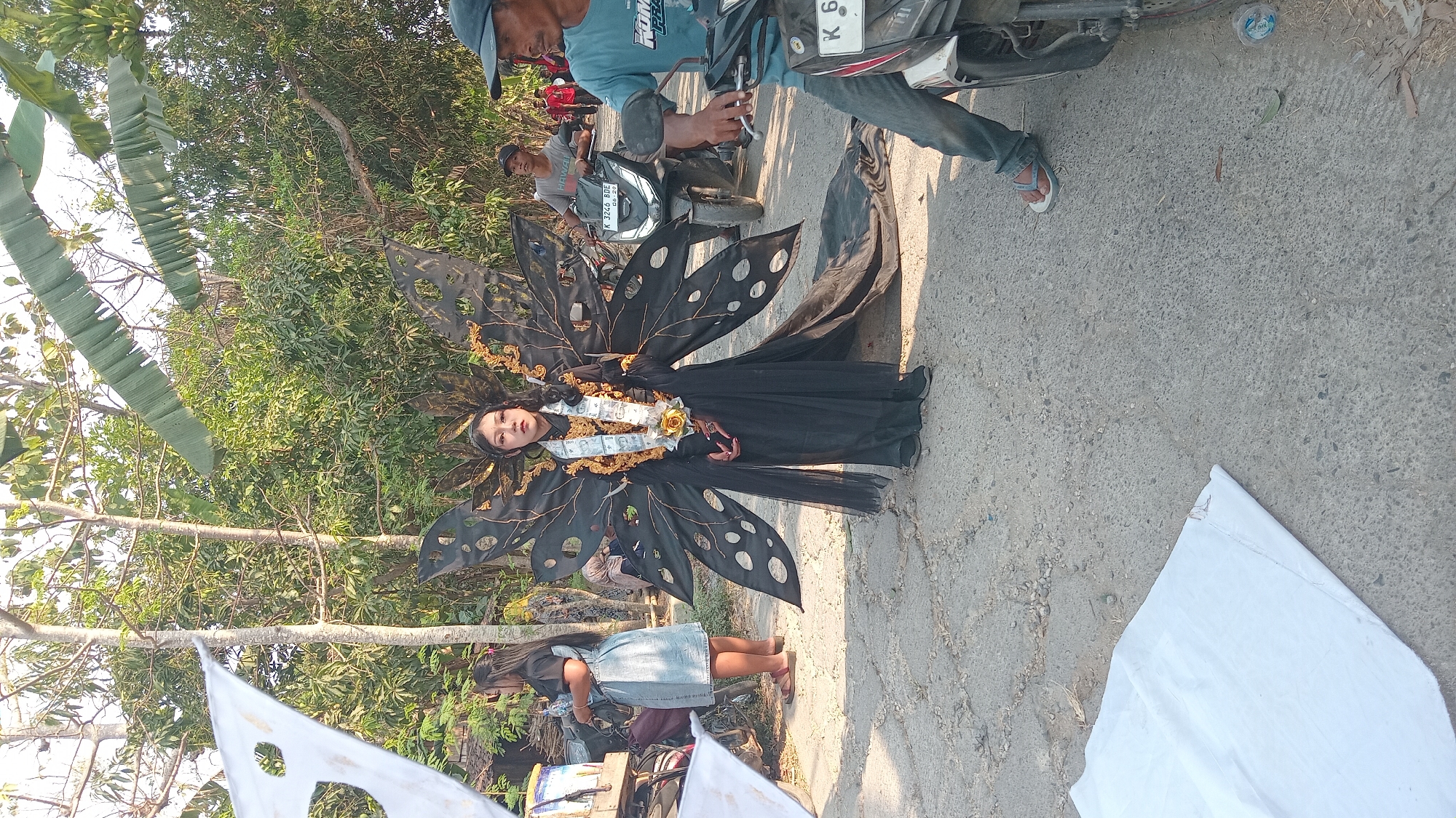 In Kedungwaru, Indonesia, a young woman is posing in a black, flowing dress and elaborate black butterfly wings adorned with gold accents. She wears a white sash with gold detailing and a large yellow flower around her neck. She is standing on a paved road surrounded by trees and vegetation. To her left, a woman in a denim dress stands near a cart. To her right, two men on motorcycles are visible, one in a blue t-shirt and the other in a grey t-shirt. White tarps are laid out on the ground in the foreground and to the left of the frame.