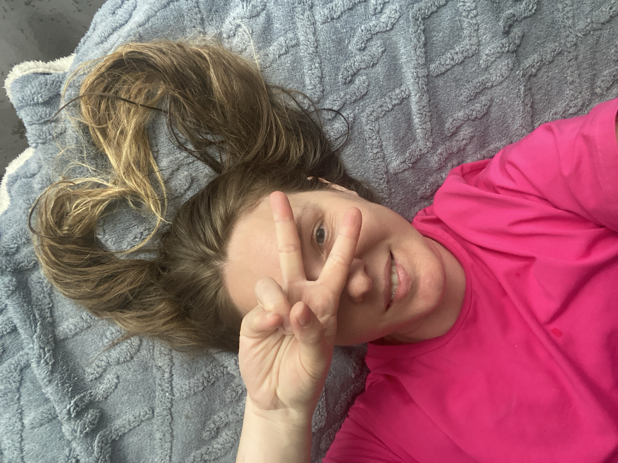 A woman is lying on a blue textured blanket. She is wearing a pink shirt and making a peace sign with her right hand. Her hair is spread out on the blanket and styled into what appears to be a heart shape. The location is not depicted in the image, so it is impossible to determine that the scene occurred in Noviy, Russia.