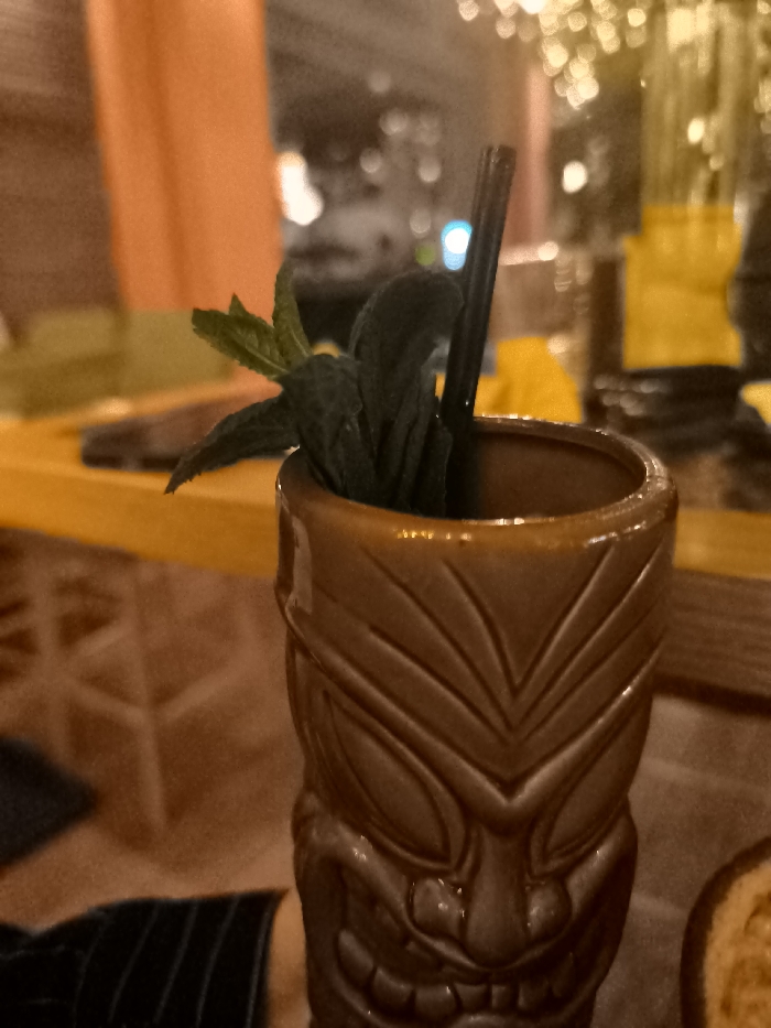 An indoor, close-up shot features a tiki mug cocktail with mint garnishes and a black straw, set against a blurred background of warm, artificial lighting. The focal point is a brown, textured ceramic tiki mug, designed with an intricate, stylized face motif visible on its lower half. Fresh green mint leaves are clustered at the top of the cocktail, protruding from the mug, alongside a slender black drinking straw.
The background is significantly out of focus, suggesting a shallow depth of field. It shows glowing bokeh lights, predominantly in warm yellow and orange hues, with a singular brighter blue light appearing on the left side of the straw. Hints of a yellow surface, possibly a bar top or table, are visible behind the mug. To the far left, an orange-colored element, possibly a wall or curtain, is present. In the lower left corner, a person's arm, dressed in a dark fabric with subtle stripes, is partially visible, indicating human presence at the table.
The overall lighting is dim and atmospheric, suggesting the photograph was taken in the evening or at night in a bar or restaurant setting, perhaps in Palaio Faliro, Greece. There is no visible text in the image.