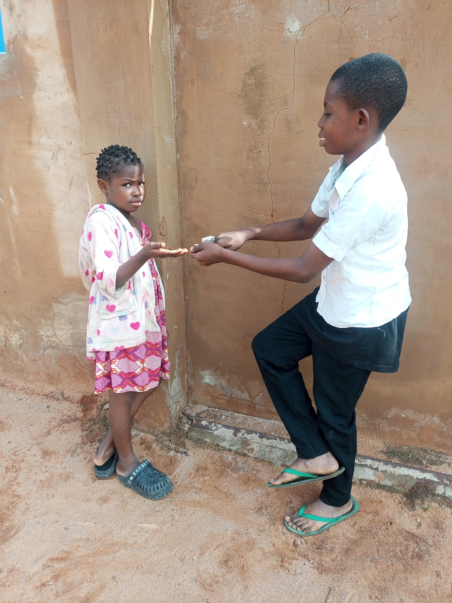 In Tempure, Nigeria, two children stand outside a building with a tan, textured wall. A younger girl on the left wears a light-colored robe with pink heart patterns over a pink dress with a geometric design. She has her left hand extended, palm up. To her right, a slightly older child in a white short-sleeved shirt and black pants leans forward, offering something small and white from their left hand to the girl's palm. Both children are barefoot, with the girl wearing black clogs and the other wearing green flip-flops. The ground beneath them is sandy and appears to be dirt. The wall behind them shows signs of age and weathering, with visible cracks.