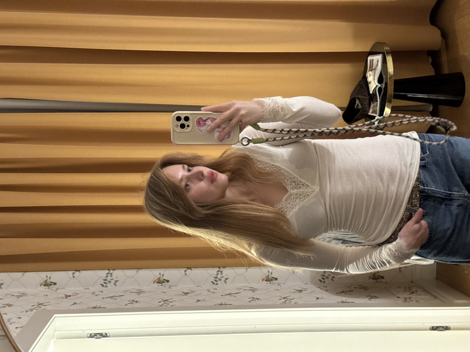 A woman takes a selfie in front of wallpapered wall and closed curtain. She wears a white long-sleeved shirt with lace details, blue jeans, and a patterned belt. Her long, light brown hair is styled with bangs. She holds a phone in her right hand, its case adorned with stickers. Behind her, a small table supports a decorative mirror and a black object. The room features a white door frame at the bottom of the frame.
