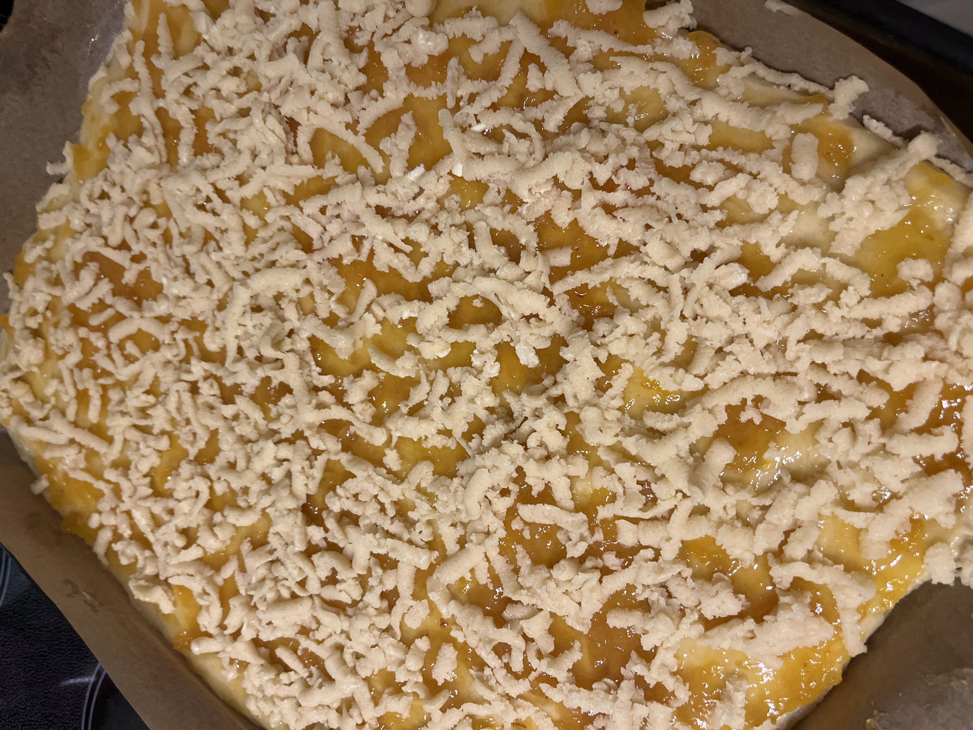 A close-up, overhead shot shows a rectangular, unbaked pastry with a golden-orange filling and a crumb topping. The pastry is on a brown baking sheet lined with parchment paper, and it appears to be in a baking pan. The filling is visible in patches between the crumb topping, which is made of small, irregular pieces of dough. The lighting suggests indoor illumination. The image does not provide any information about Kohtla-Järve linn, Estonia.