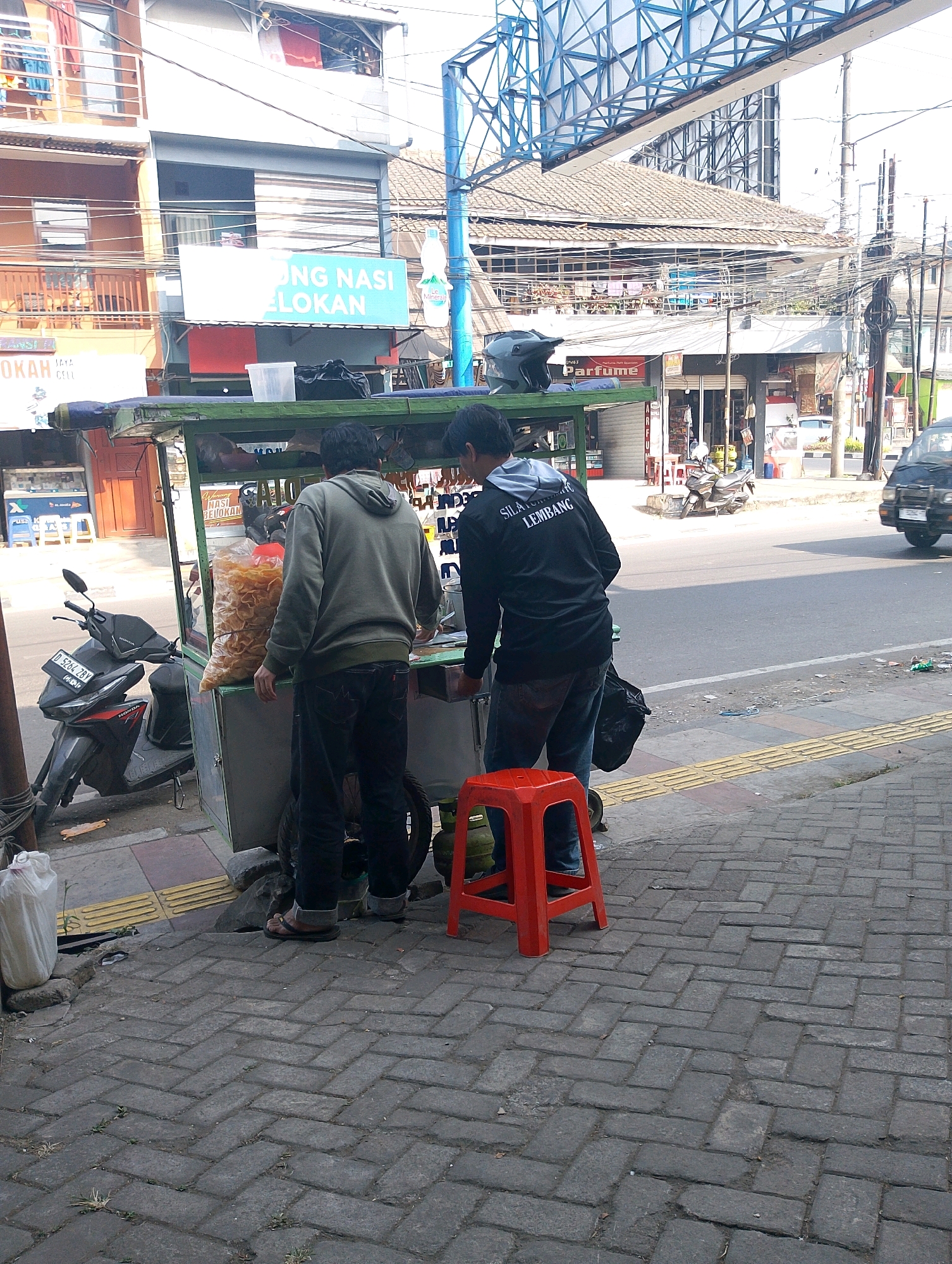 In Lembang, Indonesia, a street vendor operates a food cart on a paved sidewalk adjacent to a road. Two men stand behind the cart, one in a grey hoodie and dark pants, and the other in a black long-sleeved shirt with white text on the back. The cart displays a large bag of snacks, and a red plastic stool sits on the sidewalk. A motorcycle is parked to the left of the cart, and a black vehicle is visible on the road in the background. The scene is characterized by a mix of buildings, utility poles, and overhead wires, typical of an urban environment.