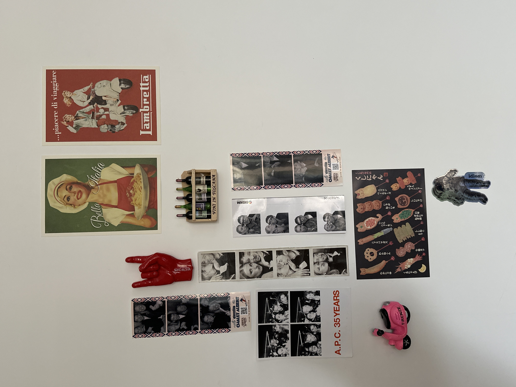 A white surface is covered with a collection of various items, mostly appearing to be magnets or stickers. Starting from the top left, there is an advertisement sticker for "Lambretta" scooters, followed by a vintage-style advertisement for "Bella Italia". Below them there is a miniature wooden wine rack containing small bottles labeled "VINI IN TOSCANA". To the right of the rack, there are black and white photo strips showing people posing, followed by a red hand with "SICILIA" written on it, stickers with Japanese food designs, a sticker of an astronaut with "idiom" written on it, and a pink moped magnet labeled "FIRENZE". There is also a sticker commemorating "A.P.C. 35 YEARS" and labels from "CHALET ALBERT".
The location is not detectable based on the image.