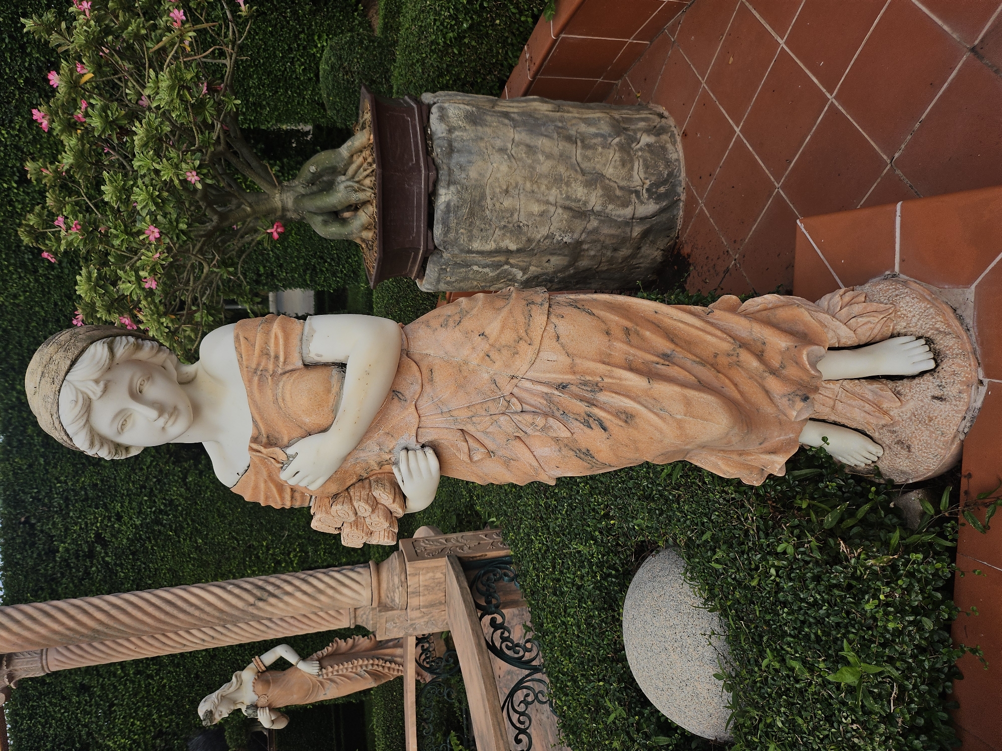 This image depicts an outdoor garden scene in Na Chom Thian, Thailand. The foreground is dominated by a life-sized marble statue of a woman in a flowing, orange-toned gown, holding a bouquet of flowers. The statue stands on a stone base, surrounded by lush green bushes and a large, smooth, gray boulder. To the left, partially visible, is another statue of a woman, also in an orange gown, positioned near a decorative stone pillar and a bench. In the background, a bonsai tree with pink flowers sits on a textured stone pedestal, set against a backdrop of meticulously trimmed green hedges. The ground is paved with reddish-brown tiles arranged in a diamond pattern. The lighting suggests it is daytime, with soft shadows cast on the ground.