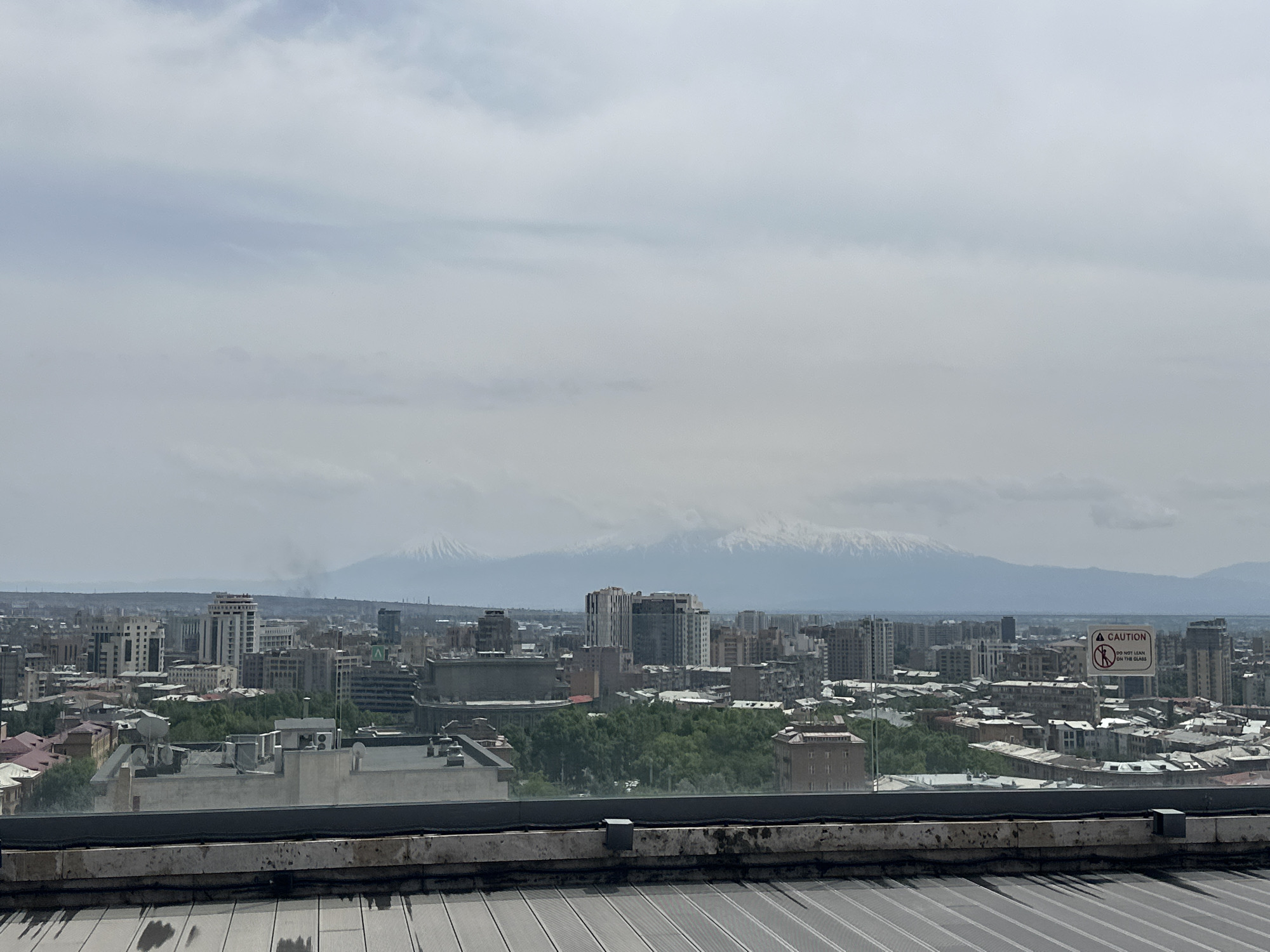 A daytime view of Yerevan, Armenia, taken from a high vantage point. The city sprawls across the midground, featuring a mix of residential and commercial buildings. A line of snow-capped mountains is visible on the horizon. A Caution sign is affixed to the glass. The view includes a flat, ribbed surface, indicating the roof of the building from which the image was captured. The sky is overcast with various cloud formations.