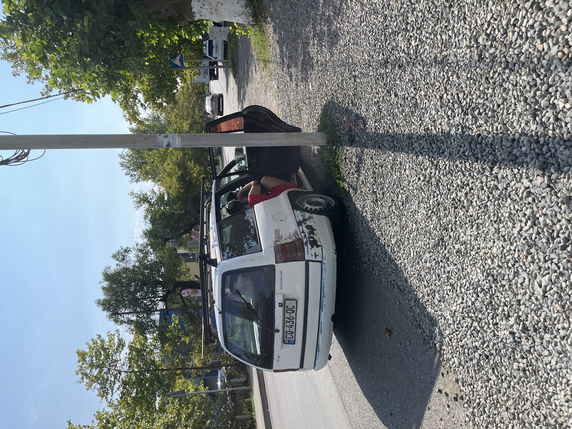 In Ureki, Georgia, a white Opel Astra station wagon is parked on the side of a street. The car's rear passenger door is open, revealing two individuals inside. A person wearing a red top is visible in the back seat, with their arm resting on the door frame. The vehicle exhibits significant damage to its rear bumper and taillights, with peeling paint and exposed metal. The license plate reads "GQ-436-QC." The street is lined with trees and buildings in the background. The foreground consists of a gravel surface, with a shadow from a utility pole cast across it. The sky is clear and blue.