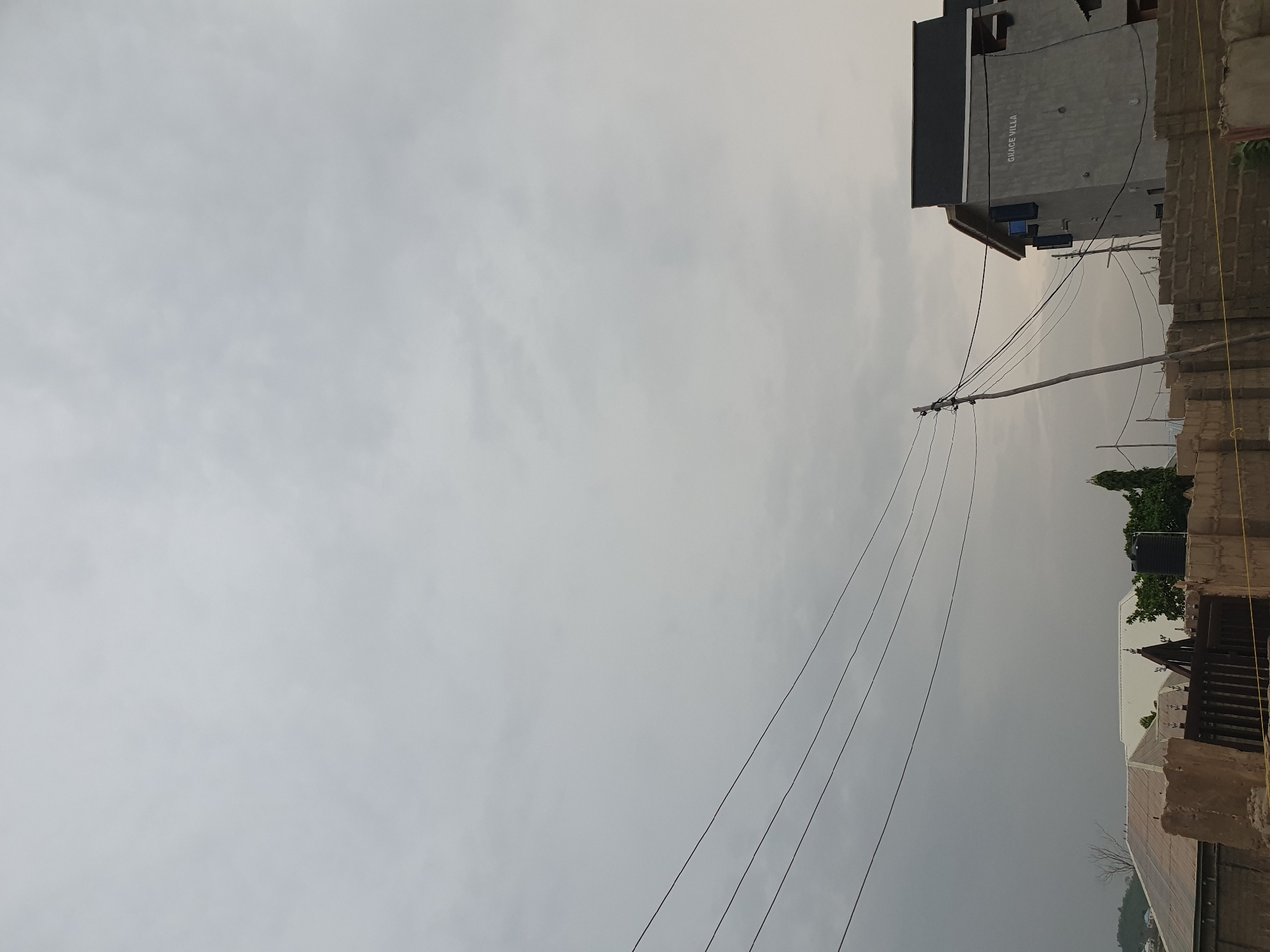 This outdoor image, taken in Lokoja, Nigeria, presents an overcast day with a grey, cloudy sky dominating the left and upper portions of the frame.
In the midground, a wooden utility pole stands upright on the right side, supporting multiple black power lines or communication cables that stretch horizontally across the image towards the left.
To the upper right, a multi-story building with grey walls and a dark, possibly sloped, roof is prominent. The text "GRACE VILLA" is clearly visible in white capital letters on the side of this building. Below its roofline, several dark, rectangular objects, likely air conditioning units, are attached to the building.
Further into the right and bottom areas of the frame, parts of other structures can be seen, including sections of a concrete or brick wall and various rooftops. A black water tank is visible on one of these distant rooftops, surrounded by some green foliage from trees or bushes. The overall scene depicts a quiet, built-up residential or mixed-use area under the muted light of a heavily clouded sky. There are no people visible, nor any discernible activities taking place.