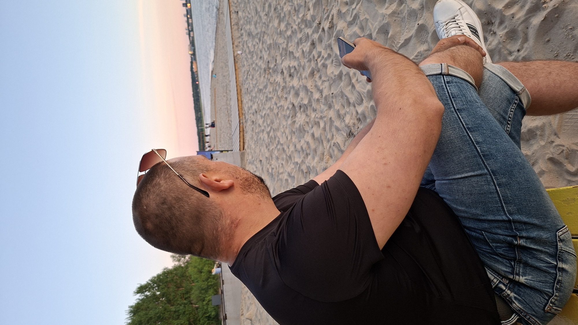 A man with short brown hair and a beard is sitting on a yellow object on a sandy beach, looking out at the water. He is wearing a black t-shirt, blue jeans, and white sneakers. Sunglasses are perched on the back of his head. He is holding a smartphone in his right hand. In the background, there is a body of water, a sandy beach with a wooden walkway, and trees. The sky is a gradient of light blue and pink, suggesting it is either sunrise or sunset. The scene appears to be at Pirita Beach in Tallinn, Estonia, given the characteristic landscape and the presence of the Pirita convent ruins in the distance, though this is not definitively visible in the crop.